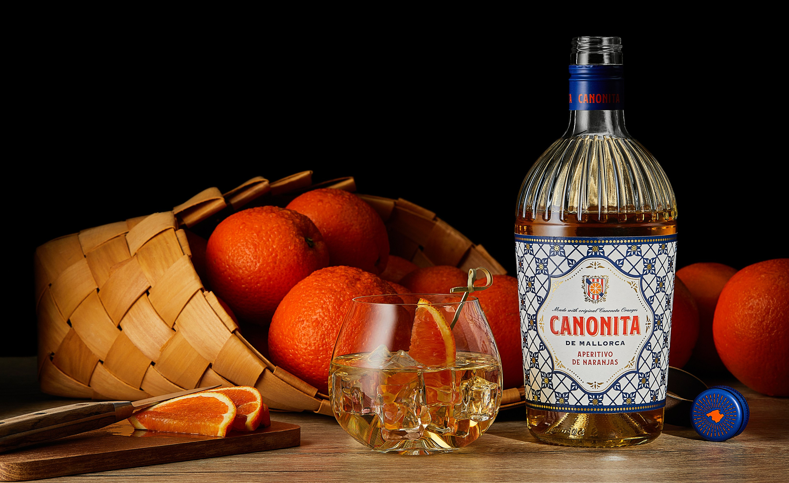 Photographer Roman Djuzev - The photograph features a bottle of Canonita de Mallorca aperitif with a striking decorative label in a Mediterranean style. The golden-hued drink is beautifully lit, highlighting the textured shape of the bottle. Beside it stands a low glass tumbler with ice and an orange slice, creating the impression of a fresh, chilled cocktail.
