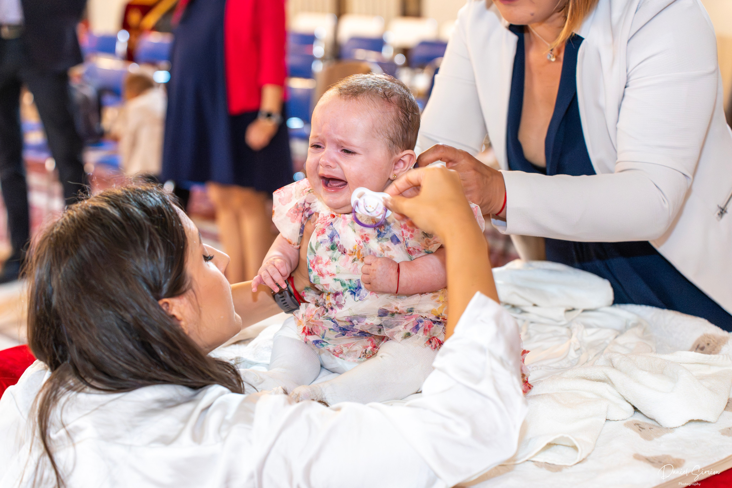 Woman comforting a crying baby wearing a floral dress indoors with blurred people in background.