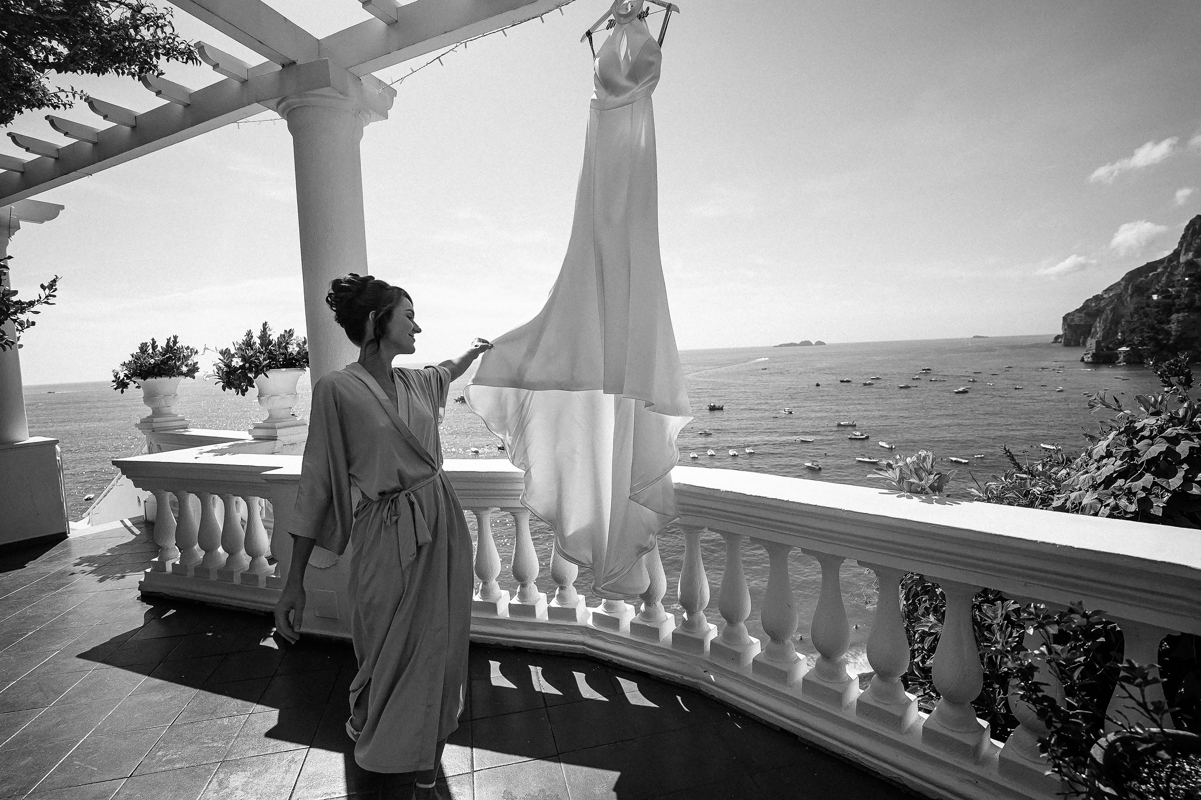 A timeless moment as the bride in a flowing lavender robe delicately reaches toward her wedding dress, with the Positano coastline and sparkling sea creating a picturesque background.