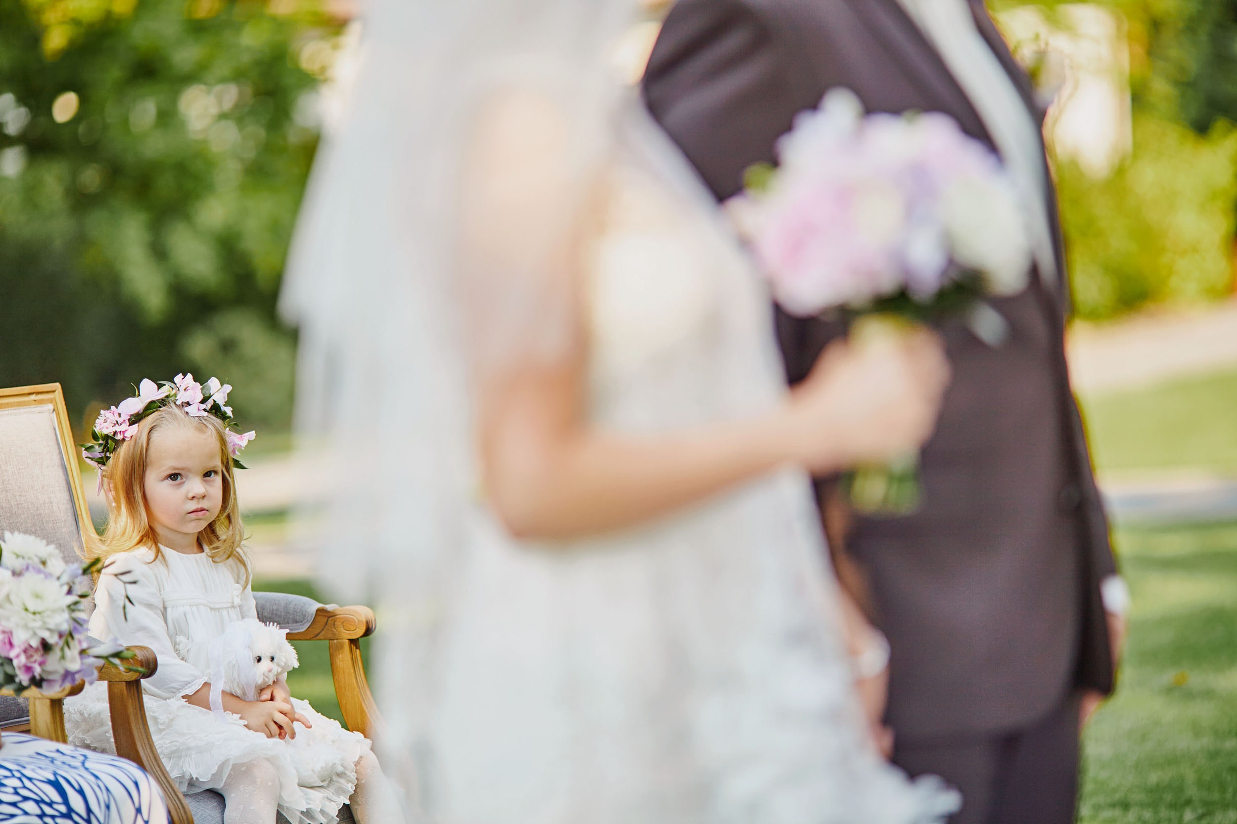 Flower girl with stuffed toy in antique chair watches bride and groom exchange vows.