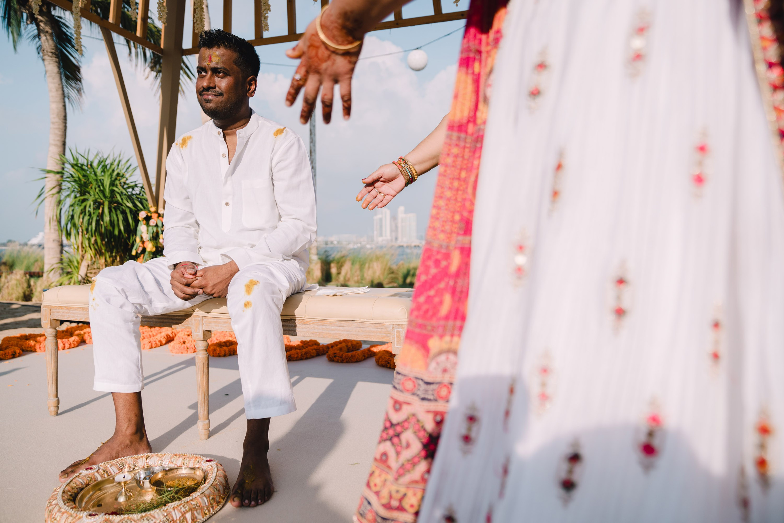 The groom is sitting on a chair and two ladies are showing at him to be ready for turmeric ceremony