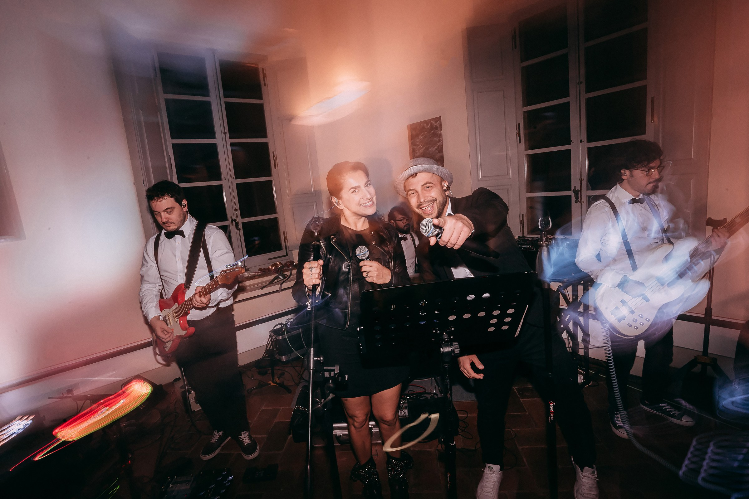 A live band performing at a wedding reception, with two vocalists center stage smiling and engaging the audience, flanked by guitarists under dramatic lighting.