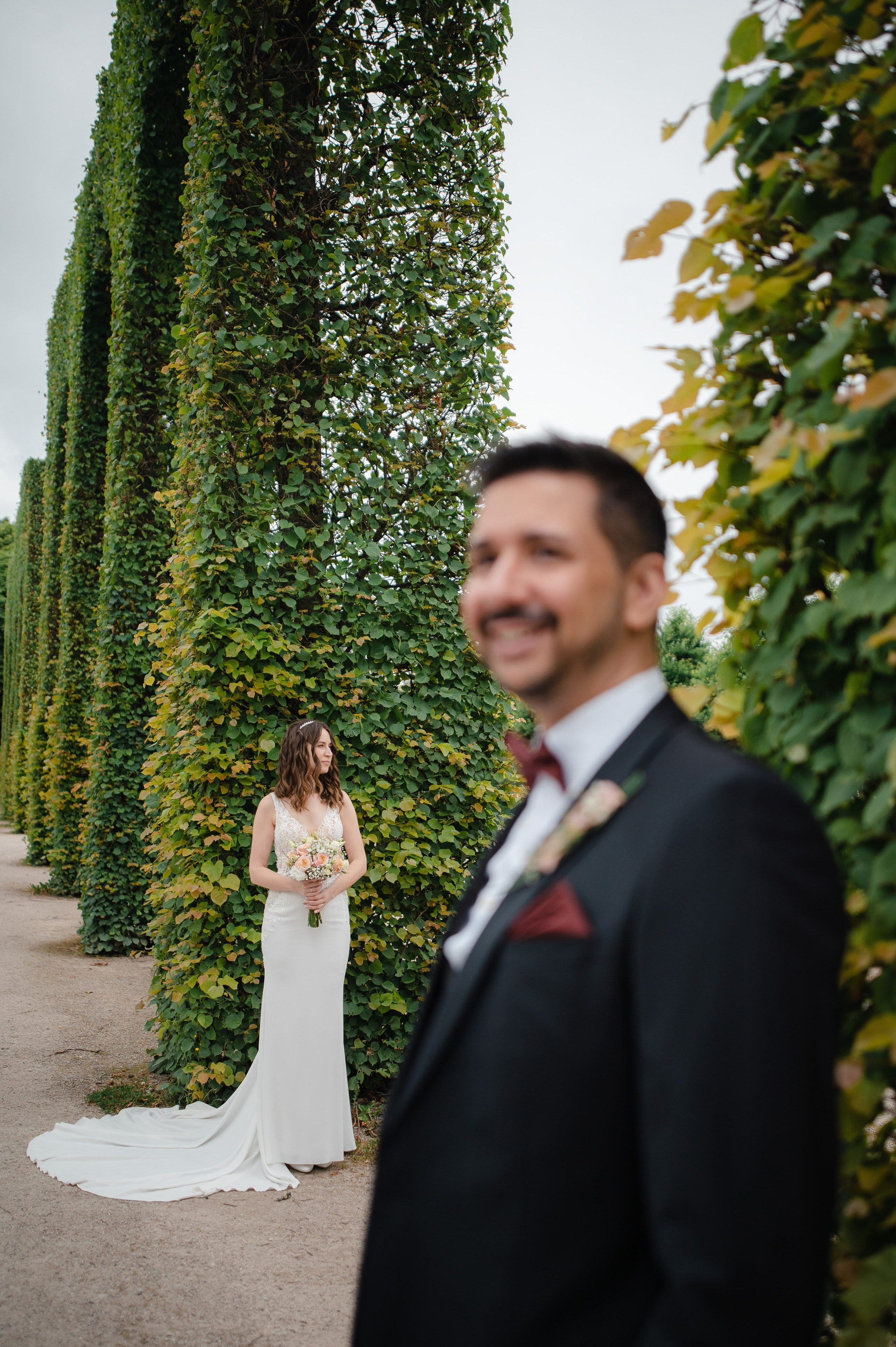 Braut mit Brautstrauß im Schlossgarten während der Hochzeitsfotos mit unscharfem Bräutigam im Vordergrund