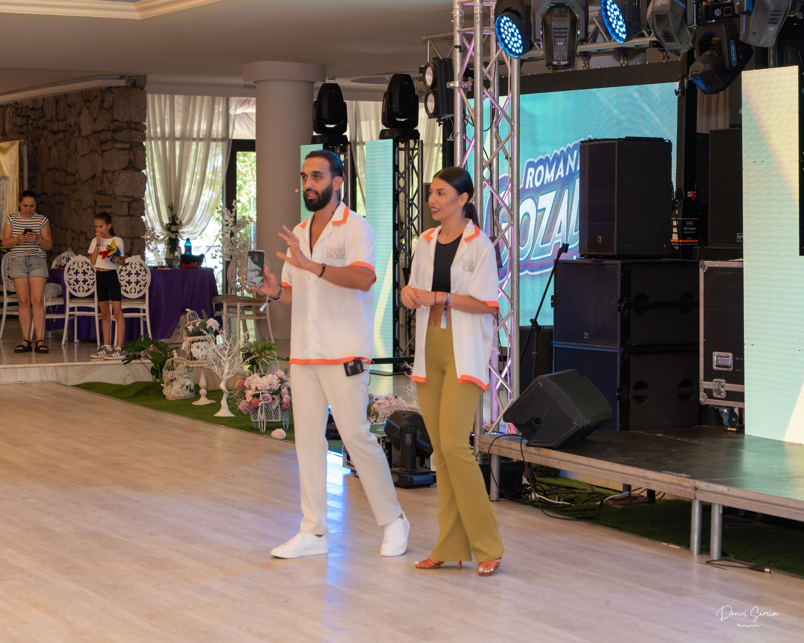 Man and woman standing on a stage speaking during an indoor event with two audience members in the background.