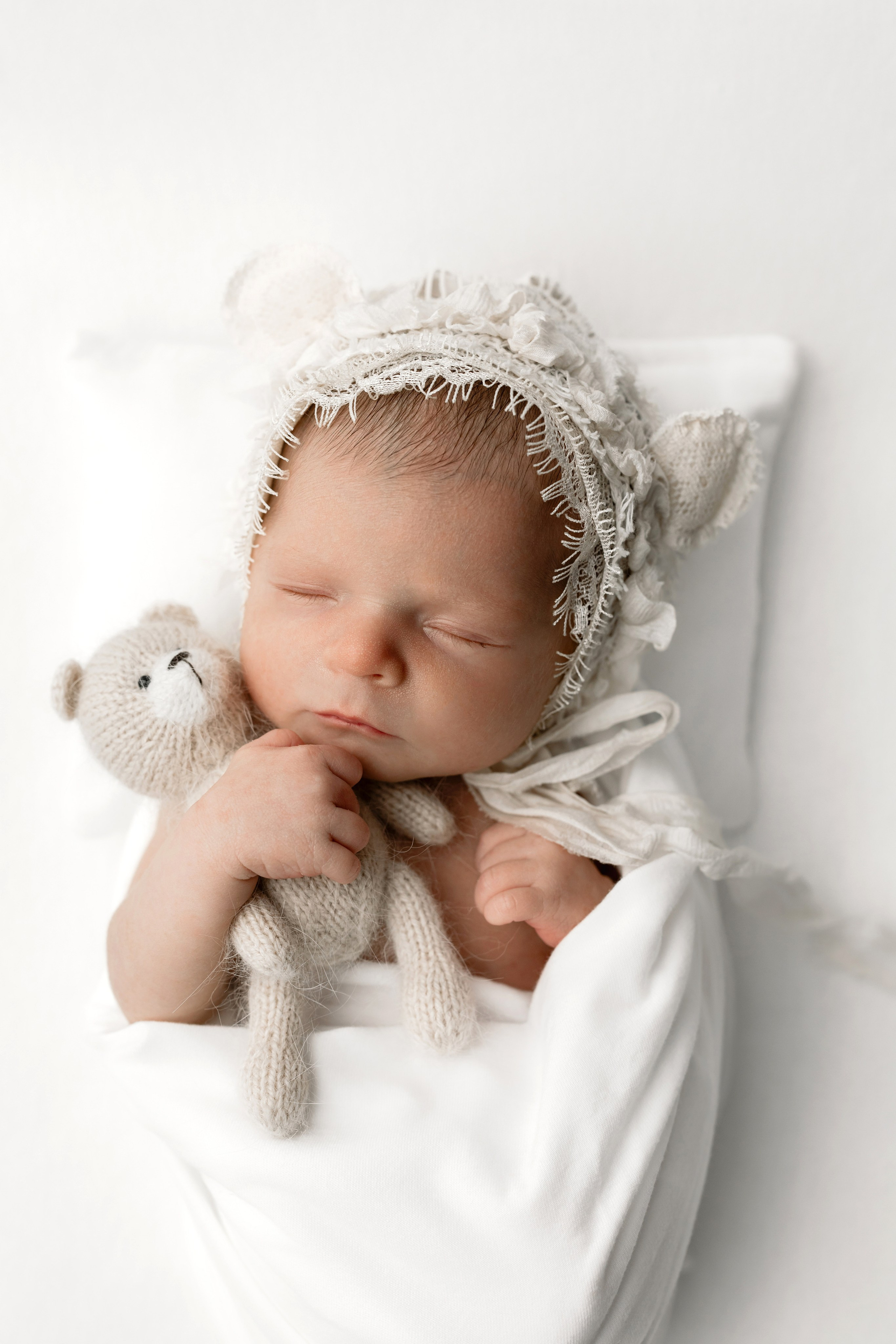 Newborn baby wrapped in a white blanket, wearing a delicate bonnet and cuddling a small teddy bear — cozy and dreamy newborn portrait in soft neutral tones.