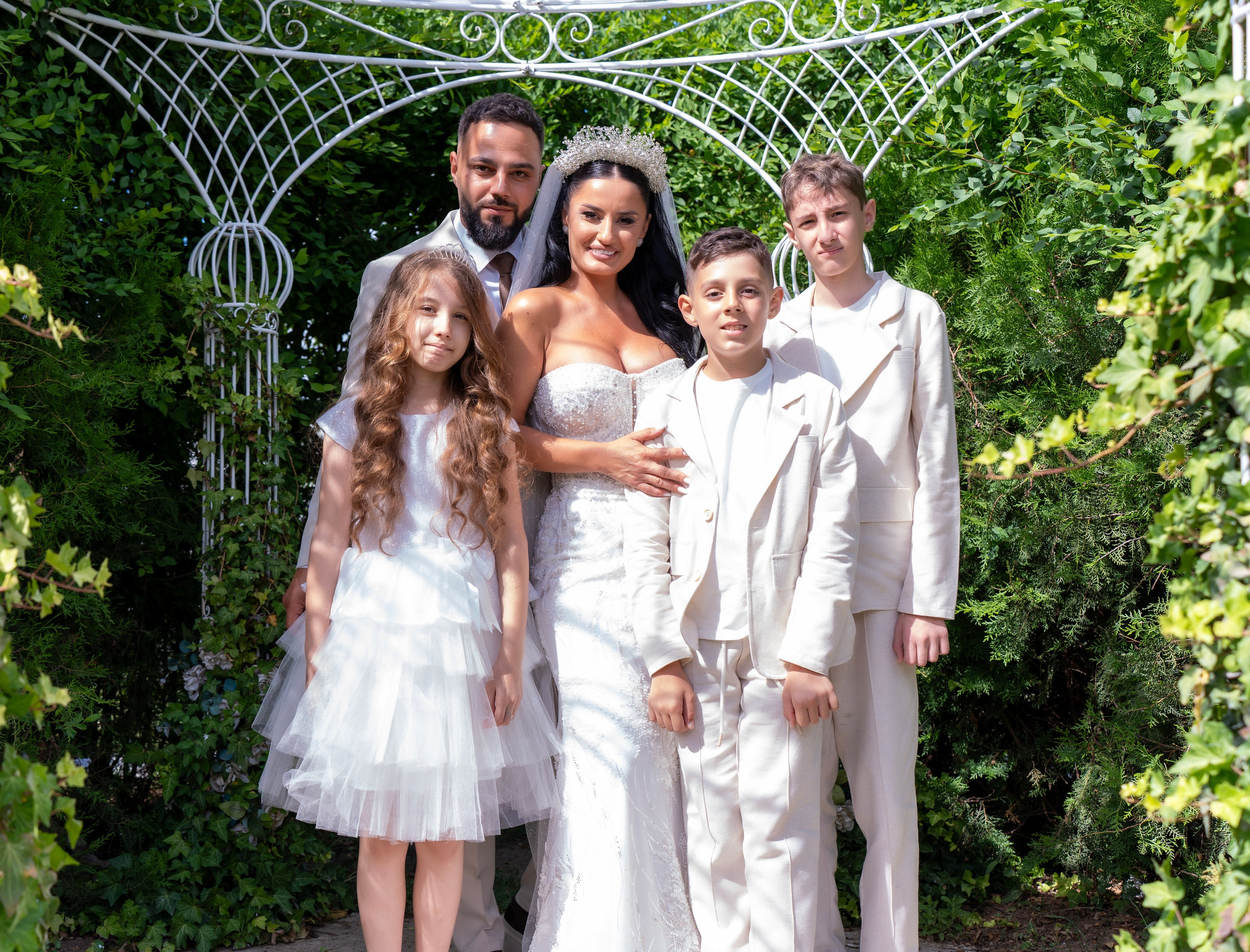 Bride and groom with three children standing under a white metal garden arch surrounded by green foliage.