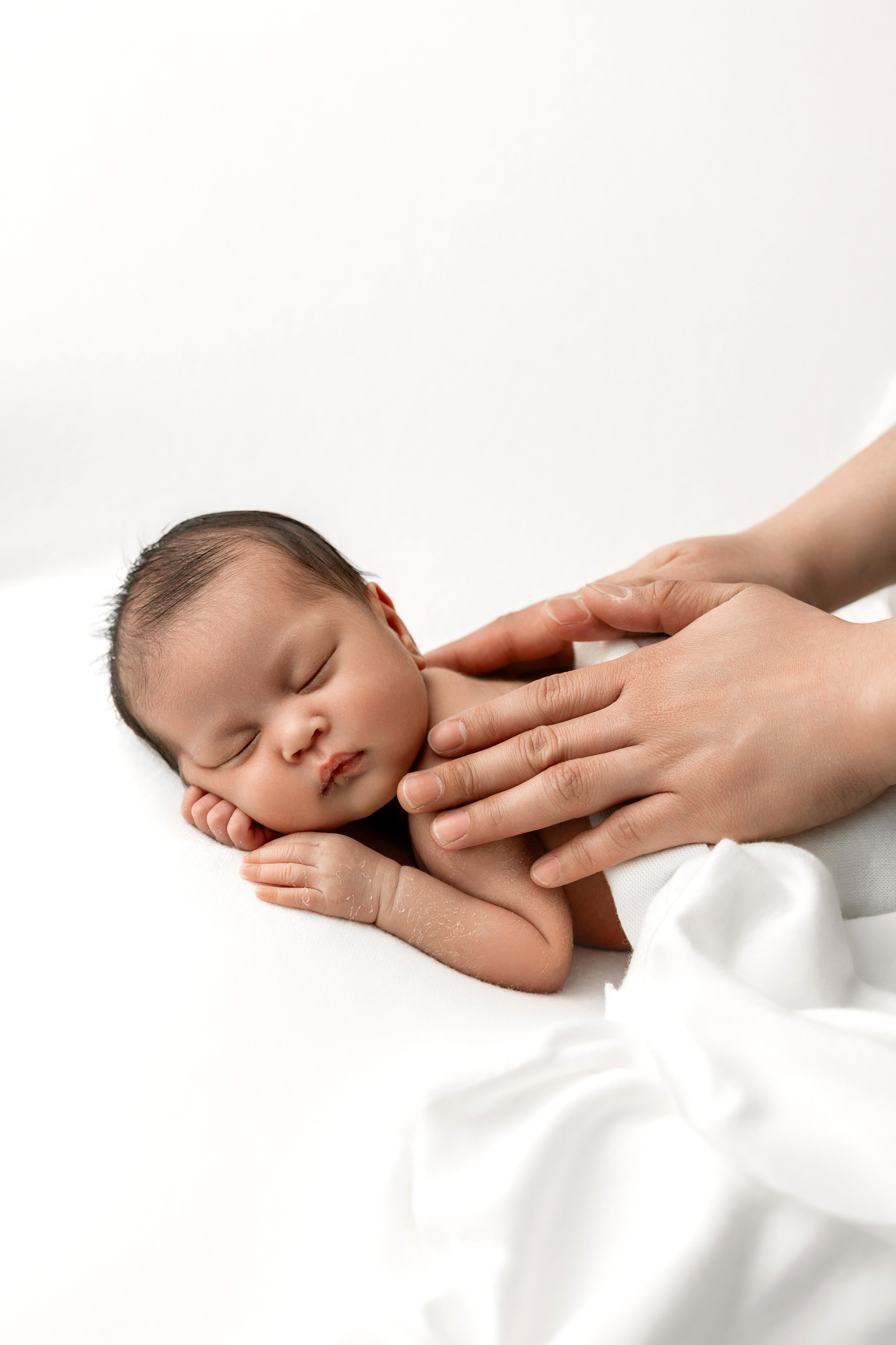 Newborn baby sleeping peacefully on a white blanket while being gently posed by hands — behind-the-scenes look at professional newborn photography session.