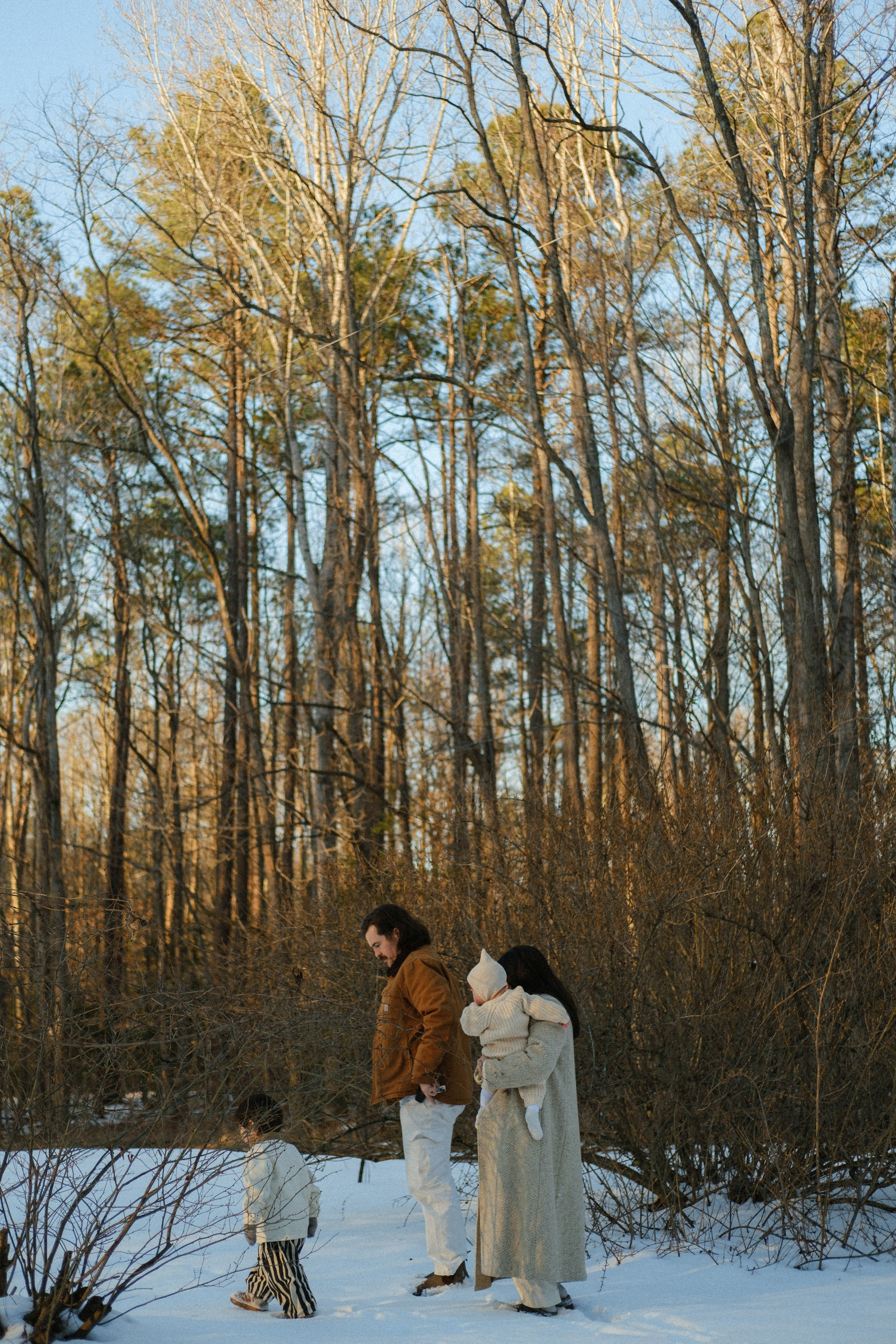 Family walking in the forest covered in snow, tall trees are above them togetehr with a blue sky, its the end of the golden hour in Richmod, VA