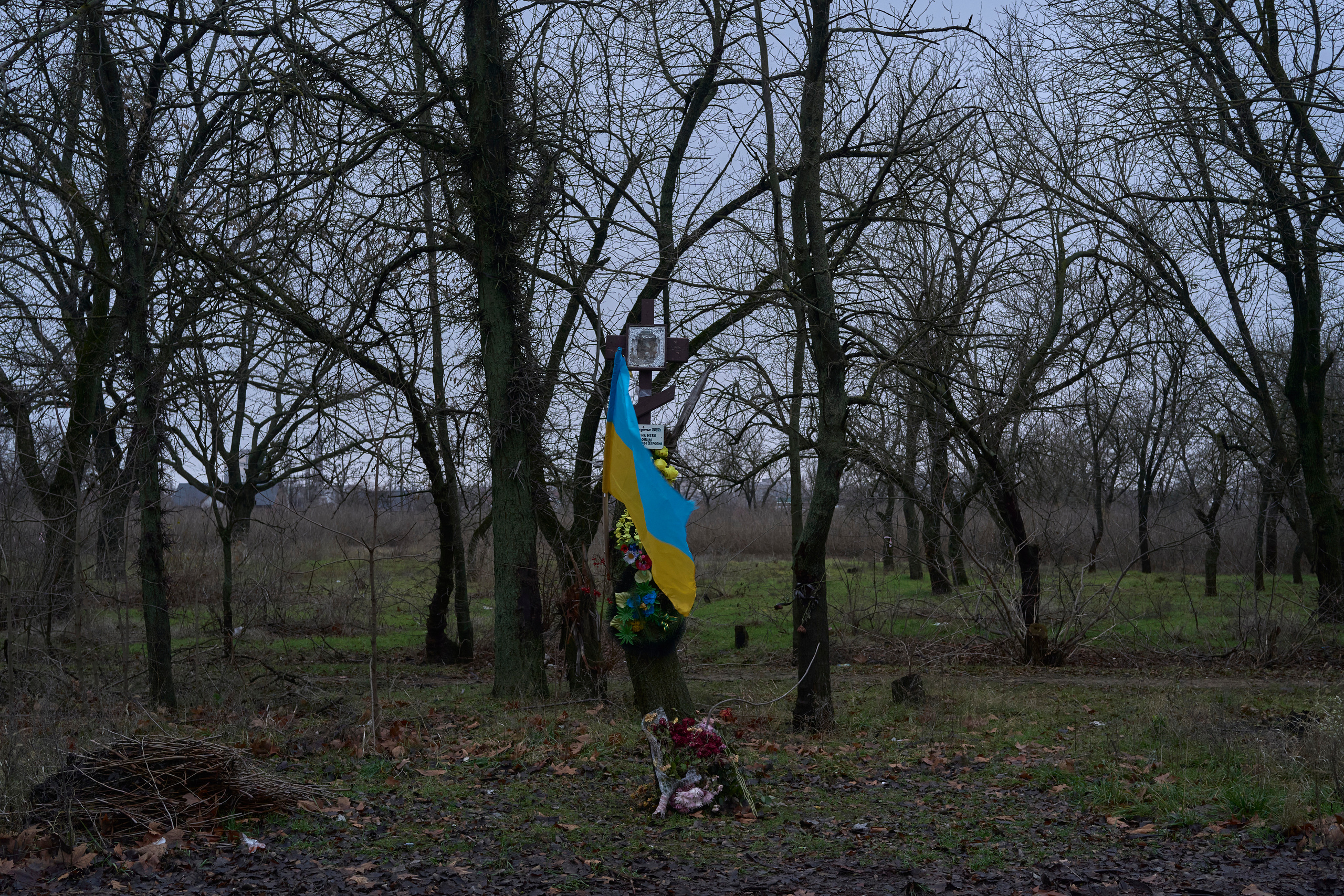 A memorial with Ukrainian flag and Orthodox cross to the fighters of territorial defence unit murdered by russians in Lilac Park. People bring funeral wreaths and flowers to the tree which was damaged by BTR bullets during the first day of entrance of russian soldiers to yhe city.