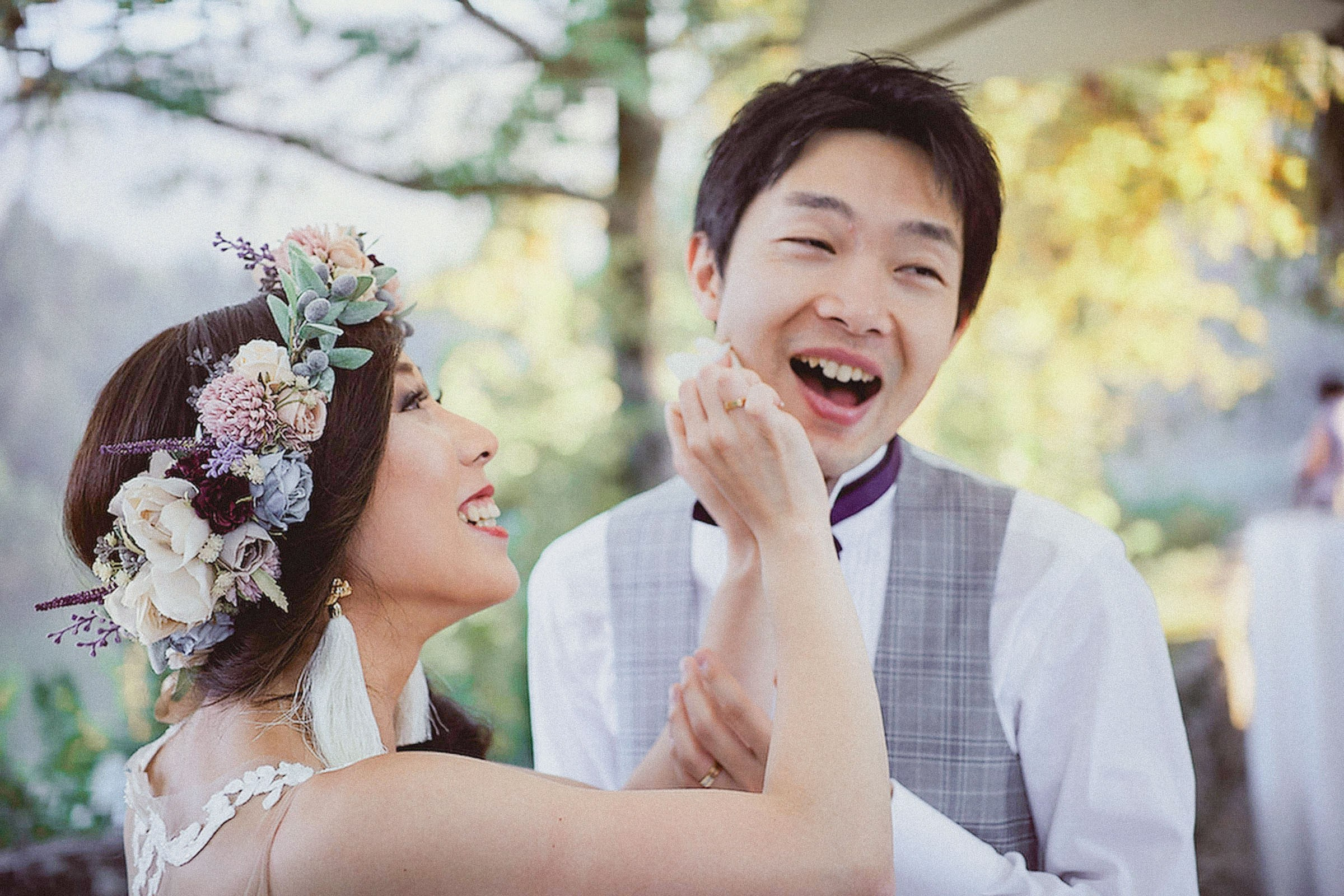 A Japanese bride, wearing a floral headpiece lovingly imitates smacking her groom's face as he mimics a pained expression.