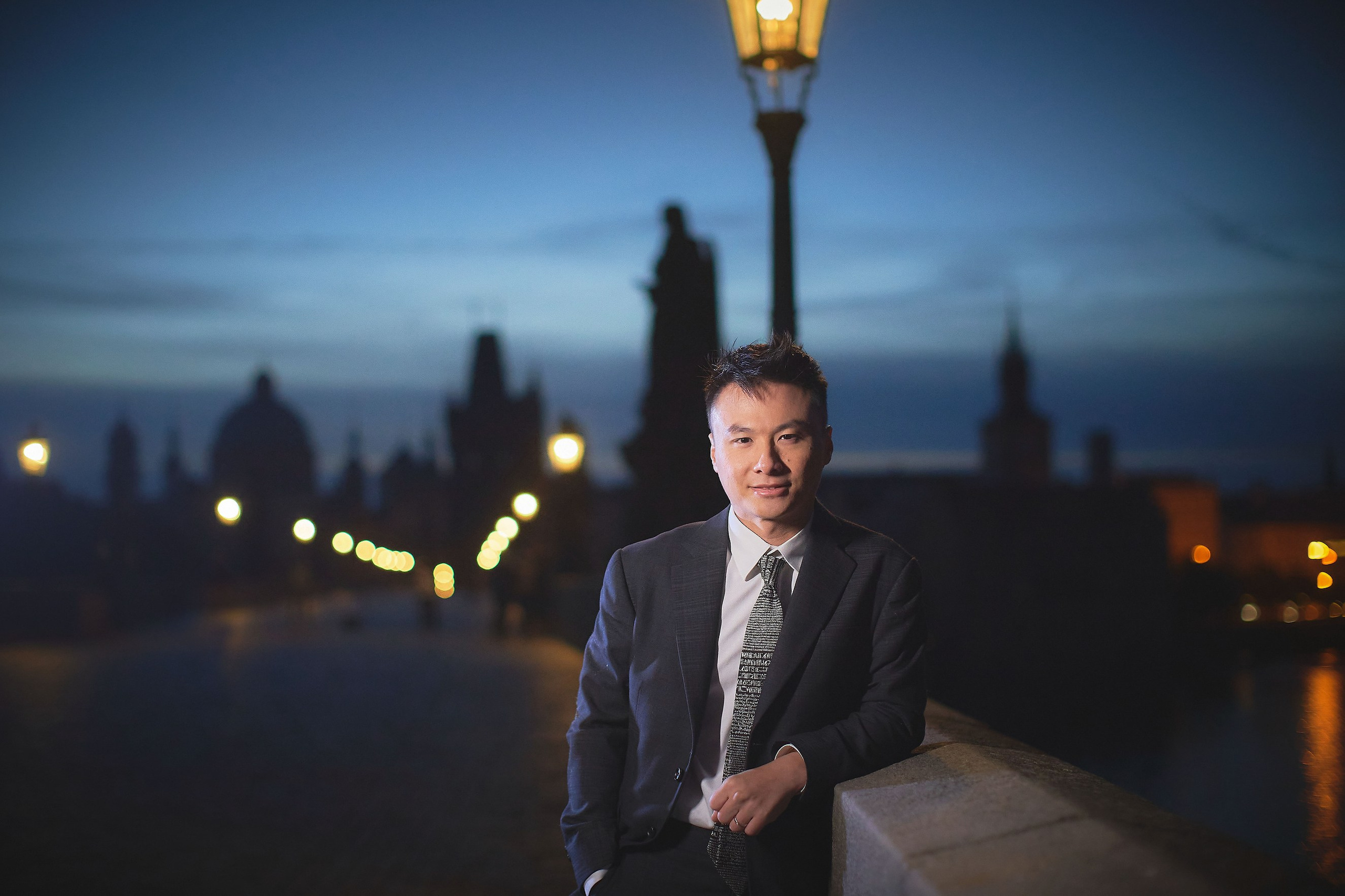 Groom Conan looking confidently at the camera with gas lamps and Charles Bridge silhouette behind.