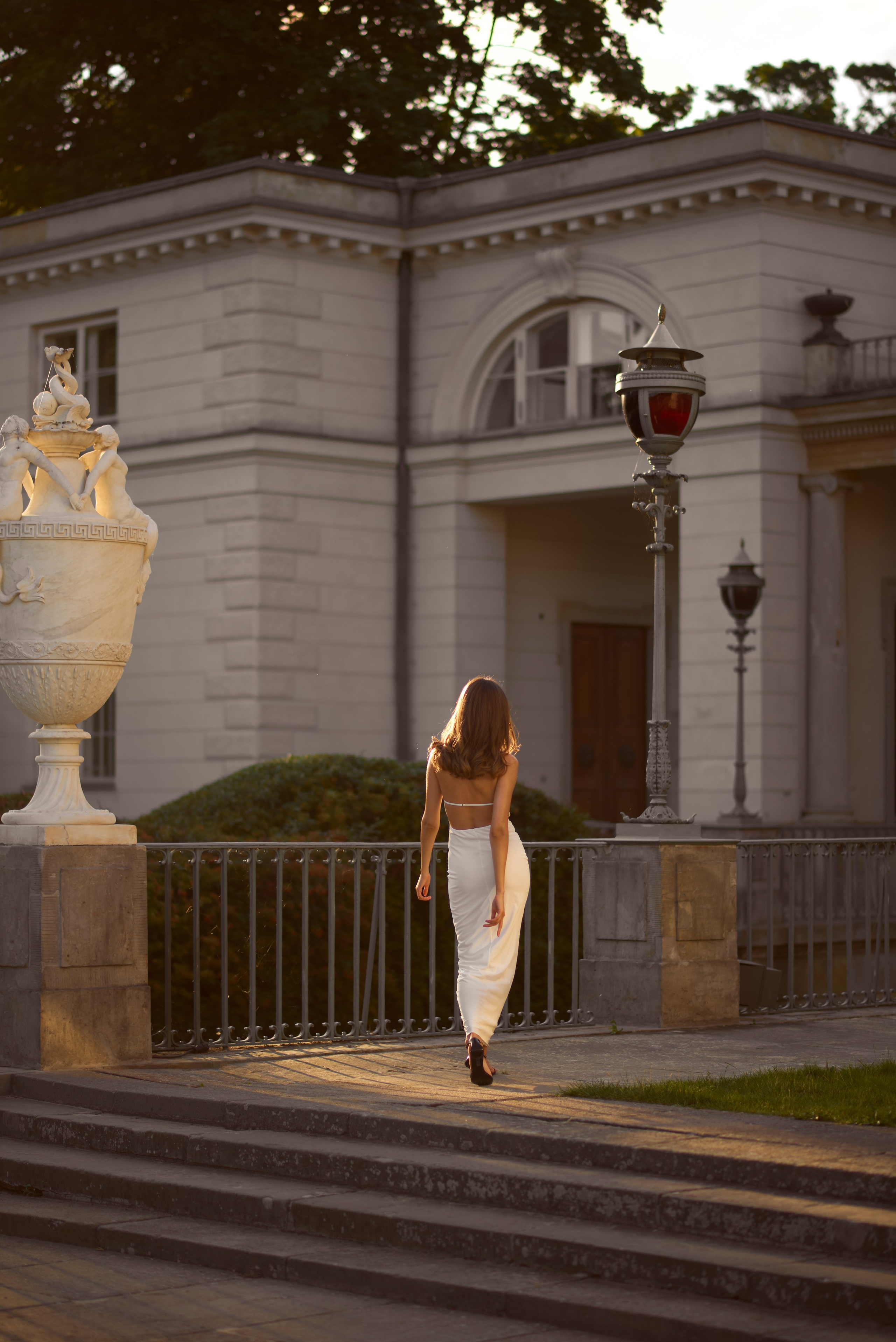 Модель у довгій білій сукні йде по терасі історичної будівлі в парку Лазенки, її силует гармонійно вписується в класицистичну архітектуру.