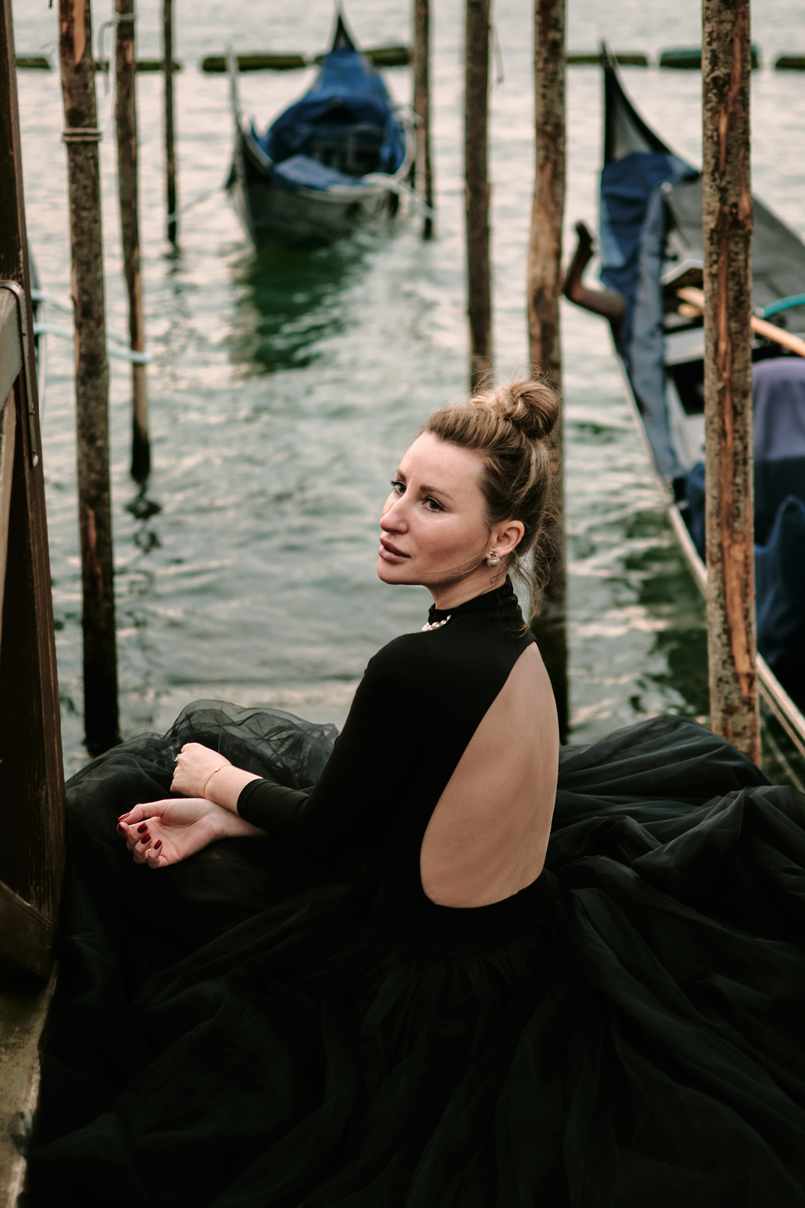 A stunning young woman in a black dress stands in San Marco Square, Venice, with the historic Arca di San Marco in the background. The architectural details of the Arca and the surrounding buildings add to the romantic atmosphere of the scene. The woman's dress drapes elegantly around her figure, adding to the captivating allure of the photo. The contrast between the dark dress and the warm colors of the surrounding buildings adds to the overall visual impact of the image, creating a sense of mystery and intrigue. Lagoon Pier in Venice. Italy