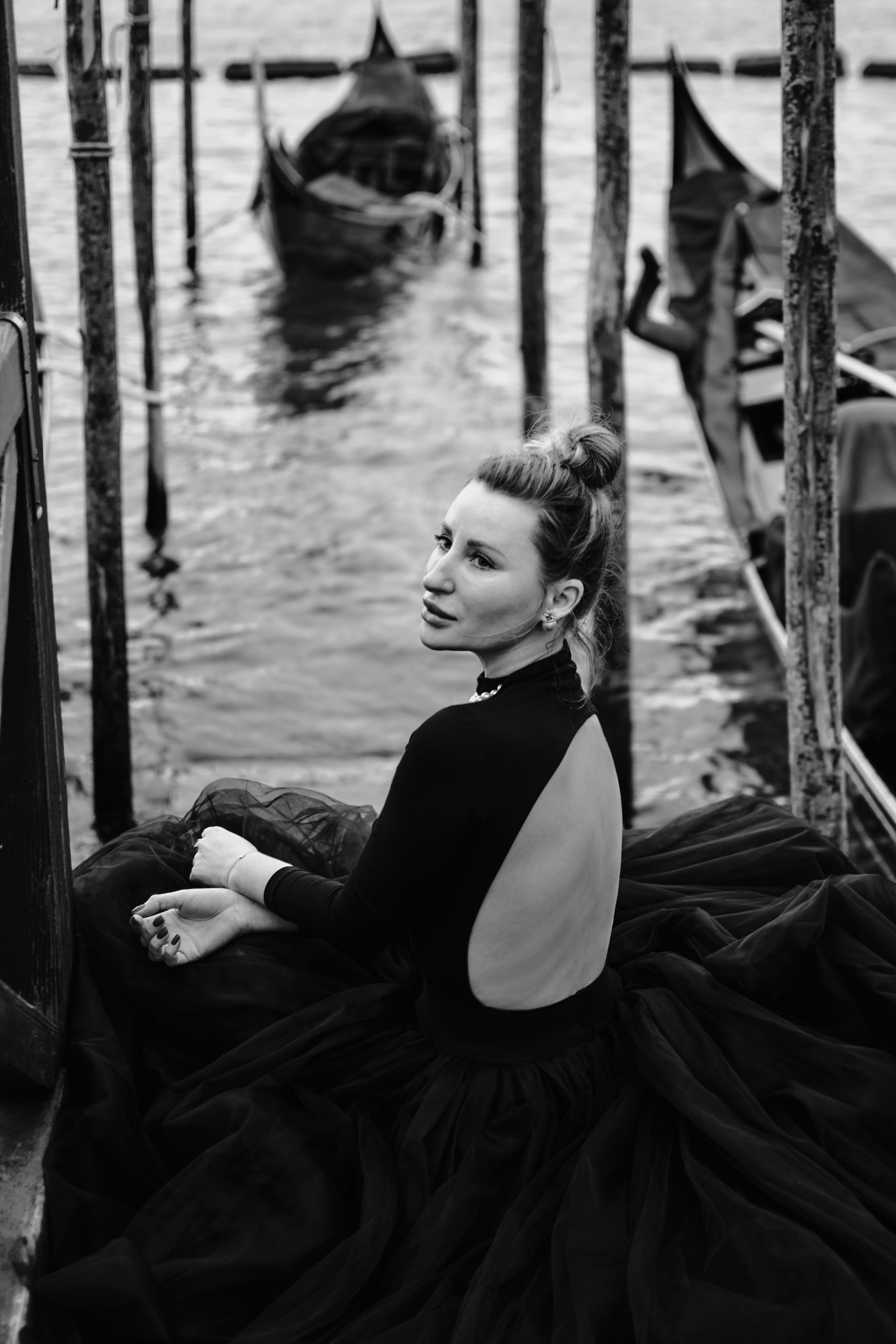 Lagoon Pier in Venice. Italy. A captivating young woman in a flowing black dress stands on a narrow bridge, framed by the intricate stonework of the arches above her. The scene is full of mystery and romance, with the gentle canal waters below and the timeless beauty of Venice's architecture surrounding her. A stunning picture of the canal in Venice with gondolas docked on the sides and a magnificent tower in the background, an iconic Instagram spot in the city.