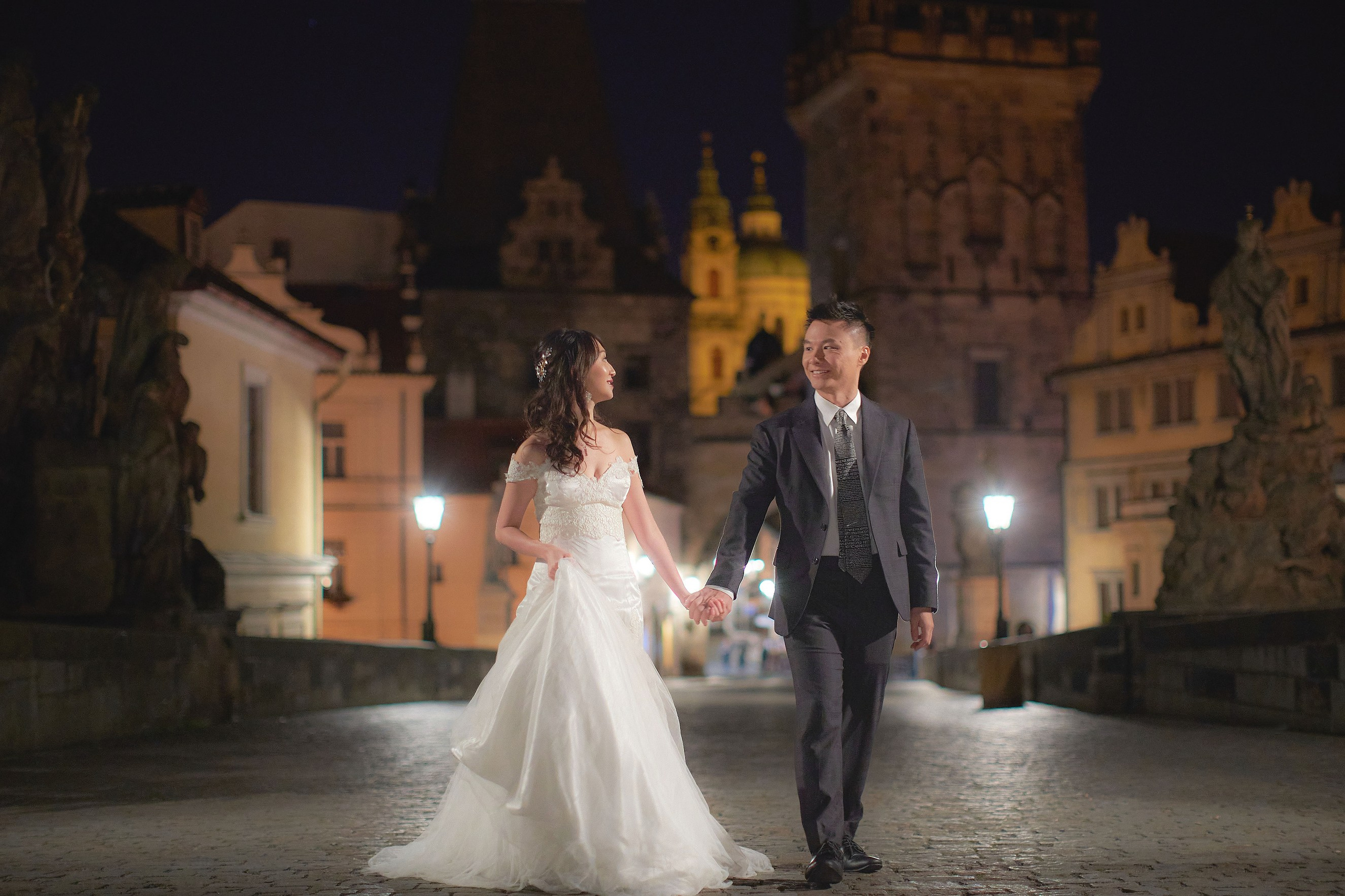 Groom Conan leading bride Eva by the hand along gas-lit Charles Bridge in early morning Prague.