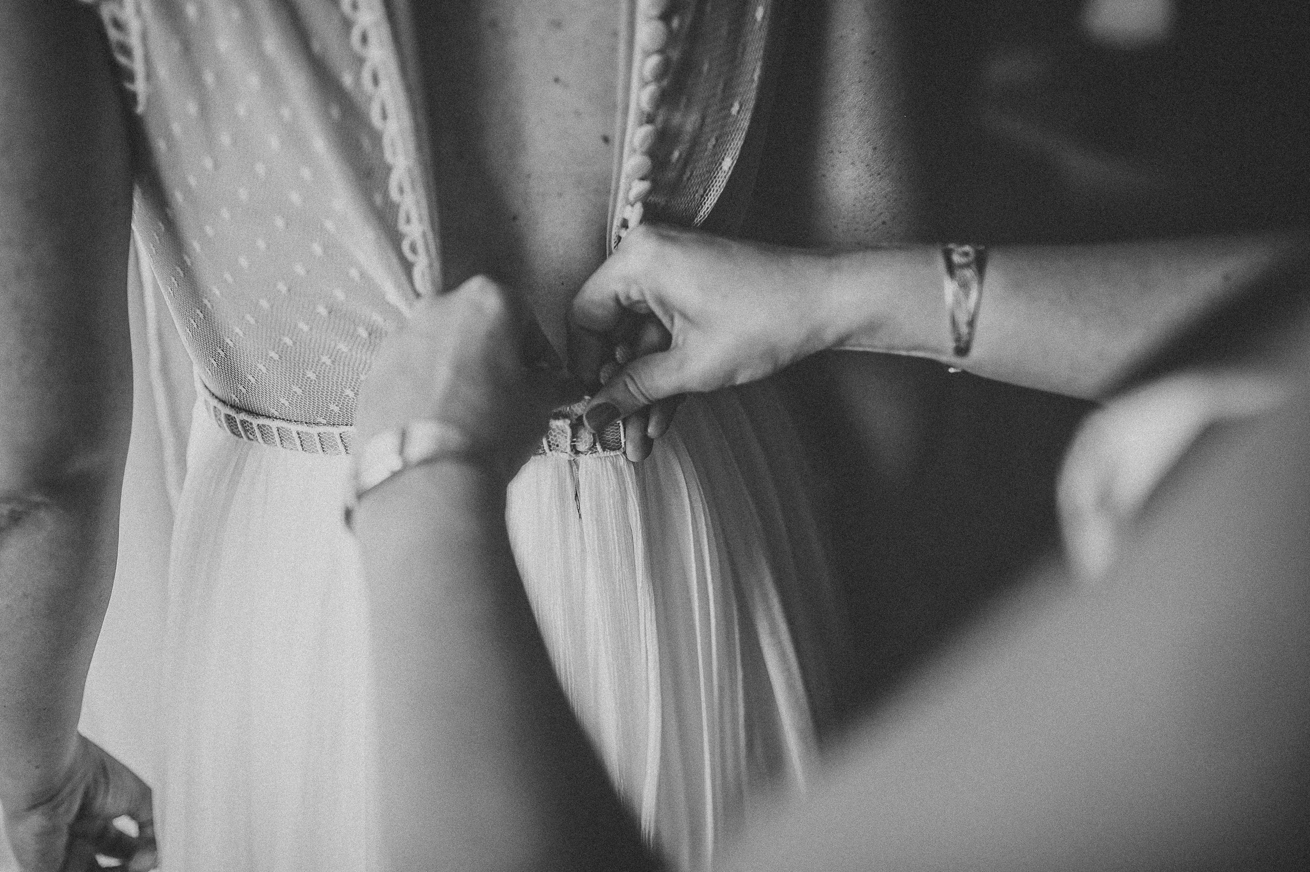 Detaliu alb-negru cu mainile nasei care inchide nasturii din spate a rochiei de miresa.