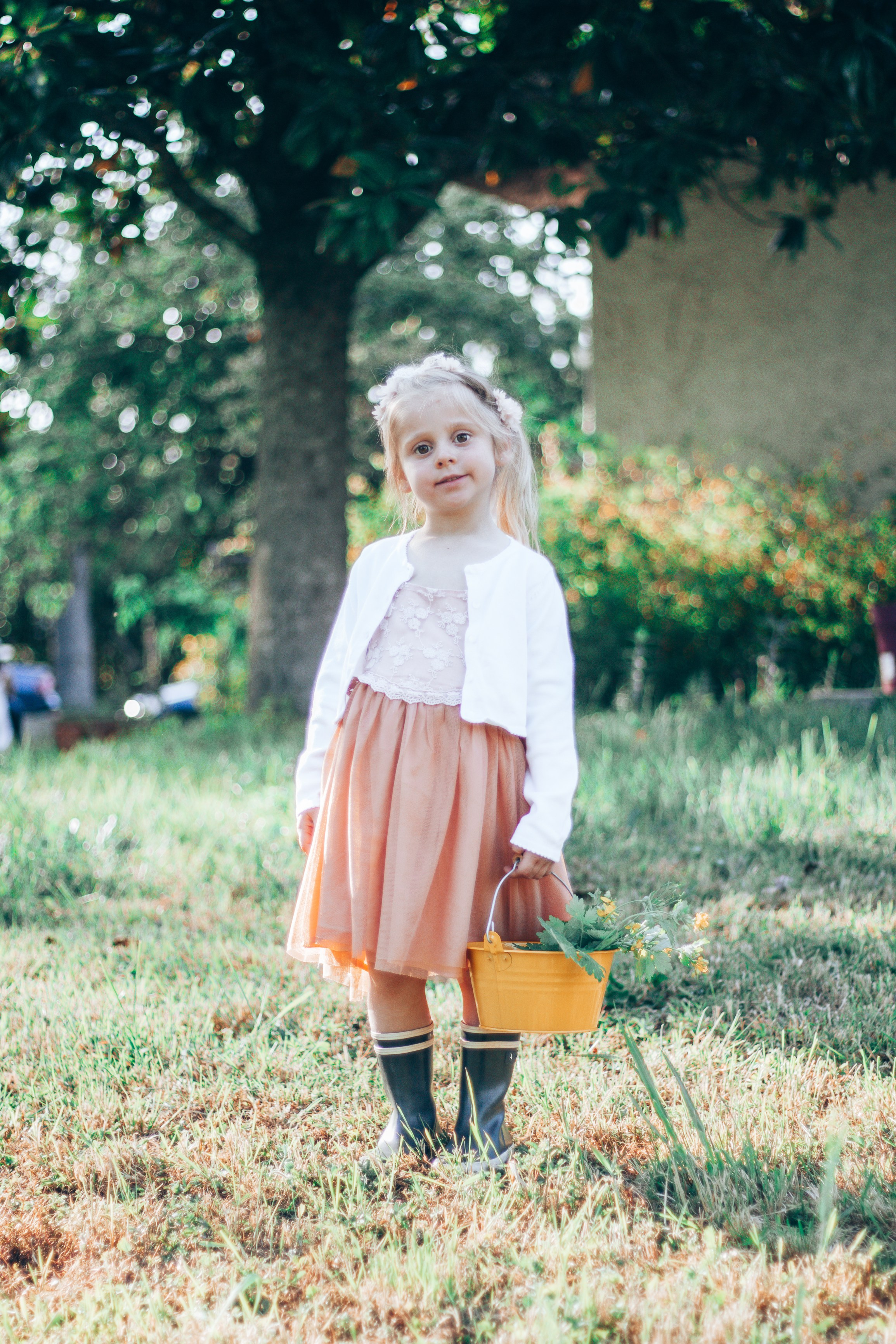 Petite fille dans un jardin à Poitiers, habillée d'une robe rose saumon et d'un gilet blanc, tenant un petit seau jaune rempli de fleurs. Scène naturelle et douce sous un grand arbre.
