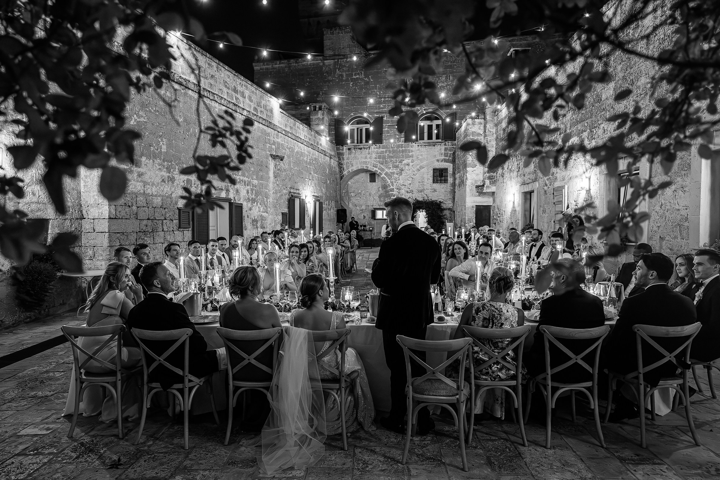 A black-and-white view of the groom giving a heartfelt toast to the seated guests during the reception, set amidst the enchanting ambiance of a candlelit courtyard.
