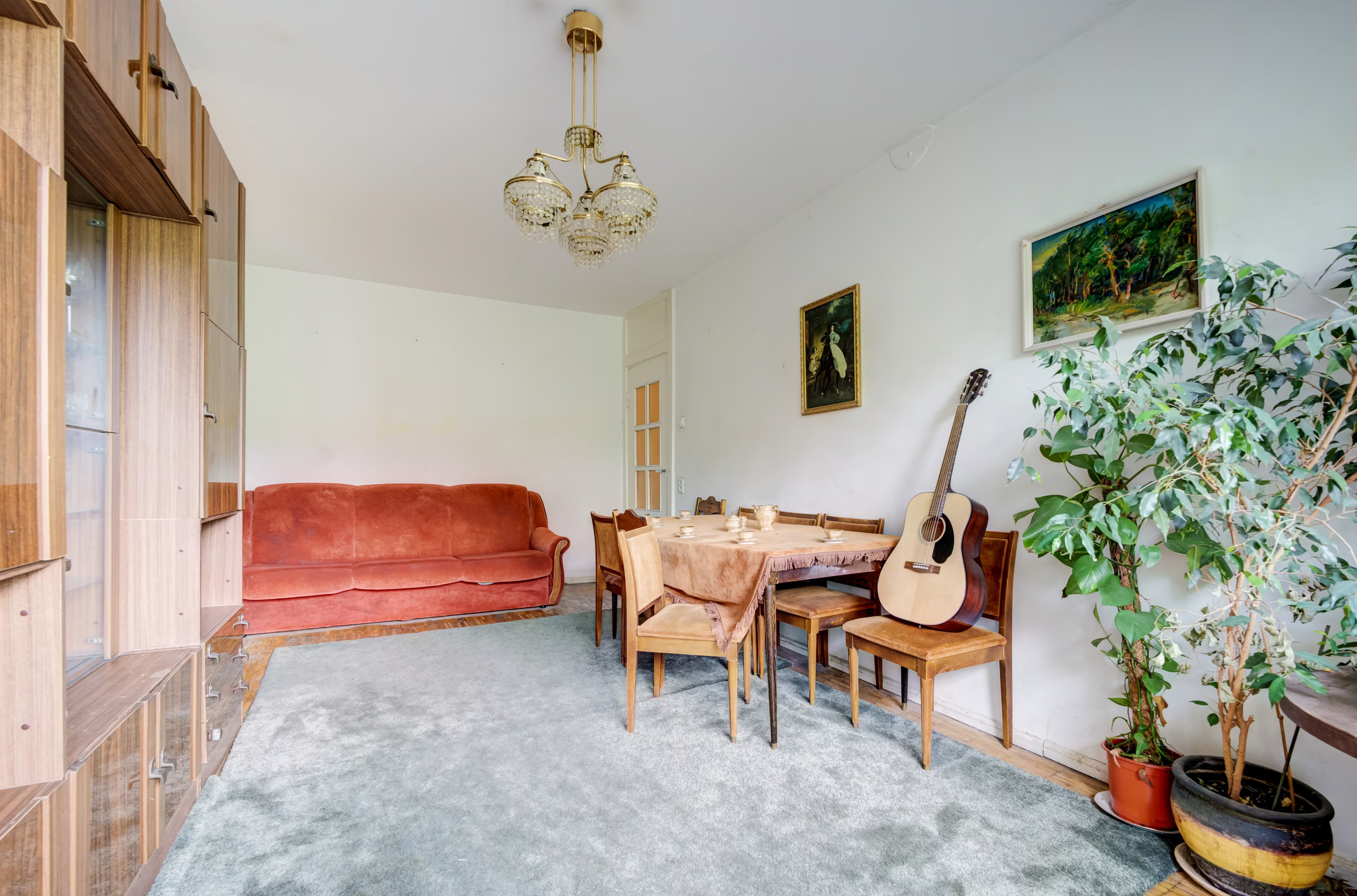 Svetainės ir valgomojo zona su raudona sofa, krištoliniu sietynu ir paveikslais ant sienų.