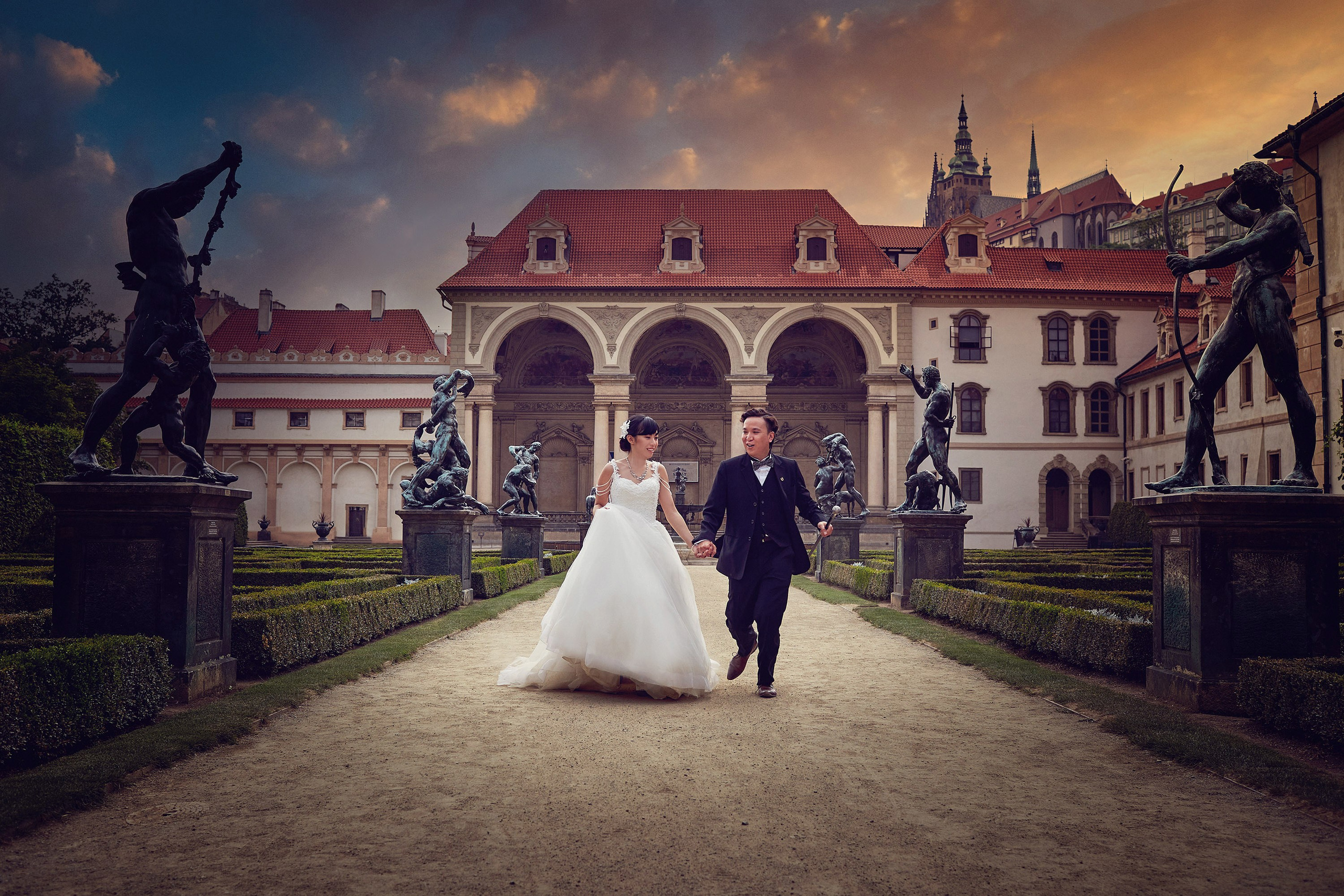 Stylish Asian bride and her groom - holding a walking cane - saunter throug the landscape of the Wallenstein Garden all alone as the setting sun flares vividly abouve Prague Castle