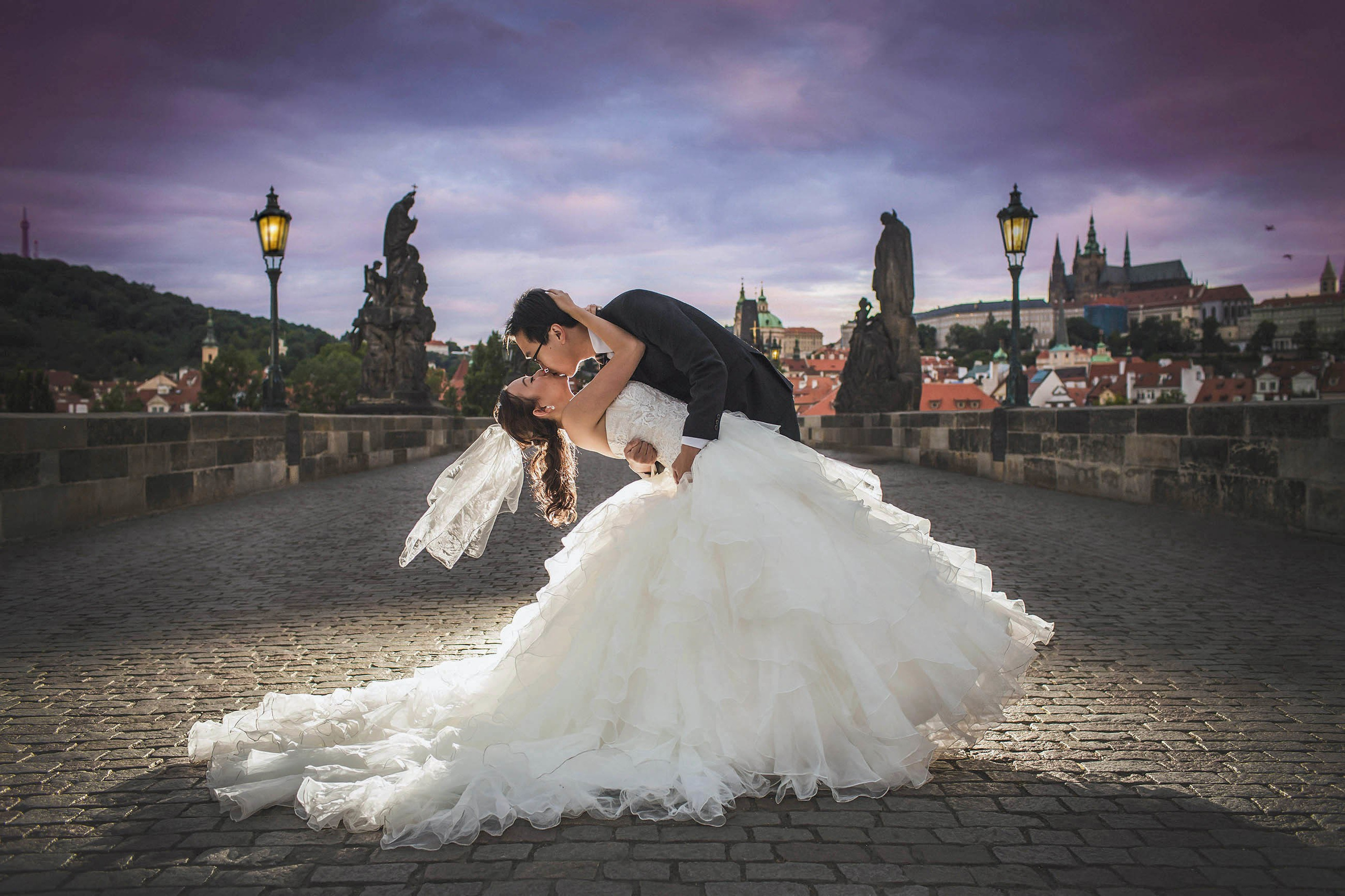 A groom dips his bride for a kiss atop the Charles Bridge under the gas lamps at night.