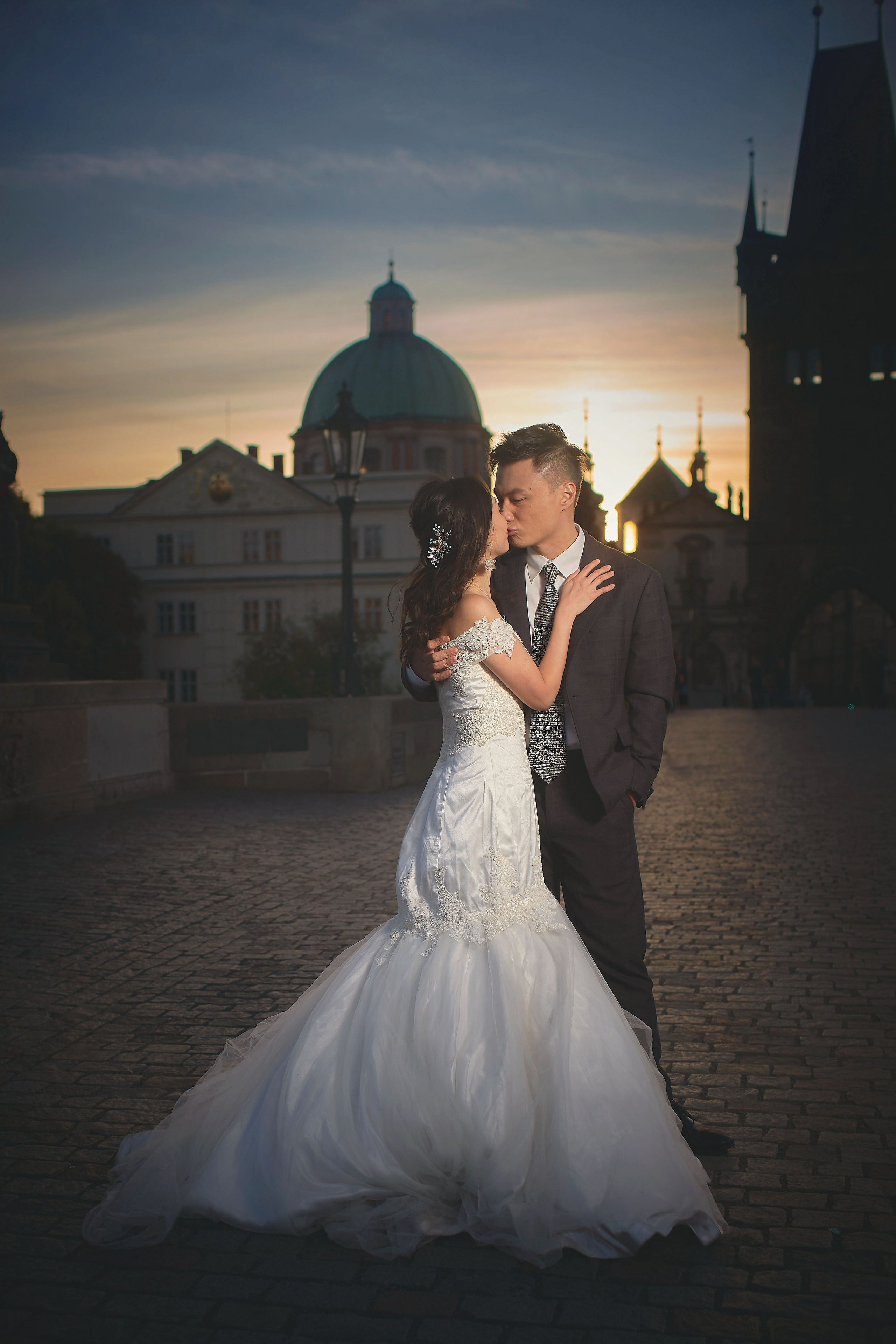 Eva and Conan share a kiss at sunrise on Charles Bridge, Prague skyline in the background.