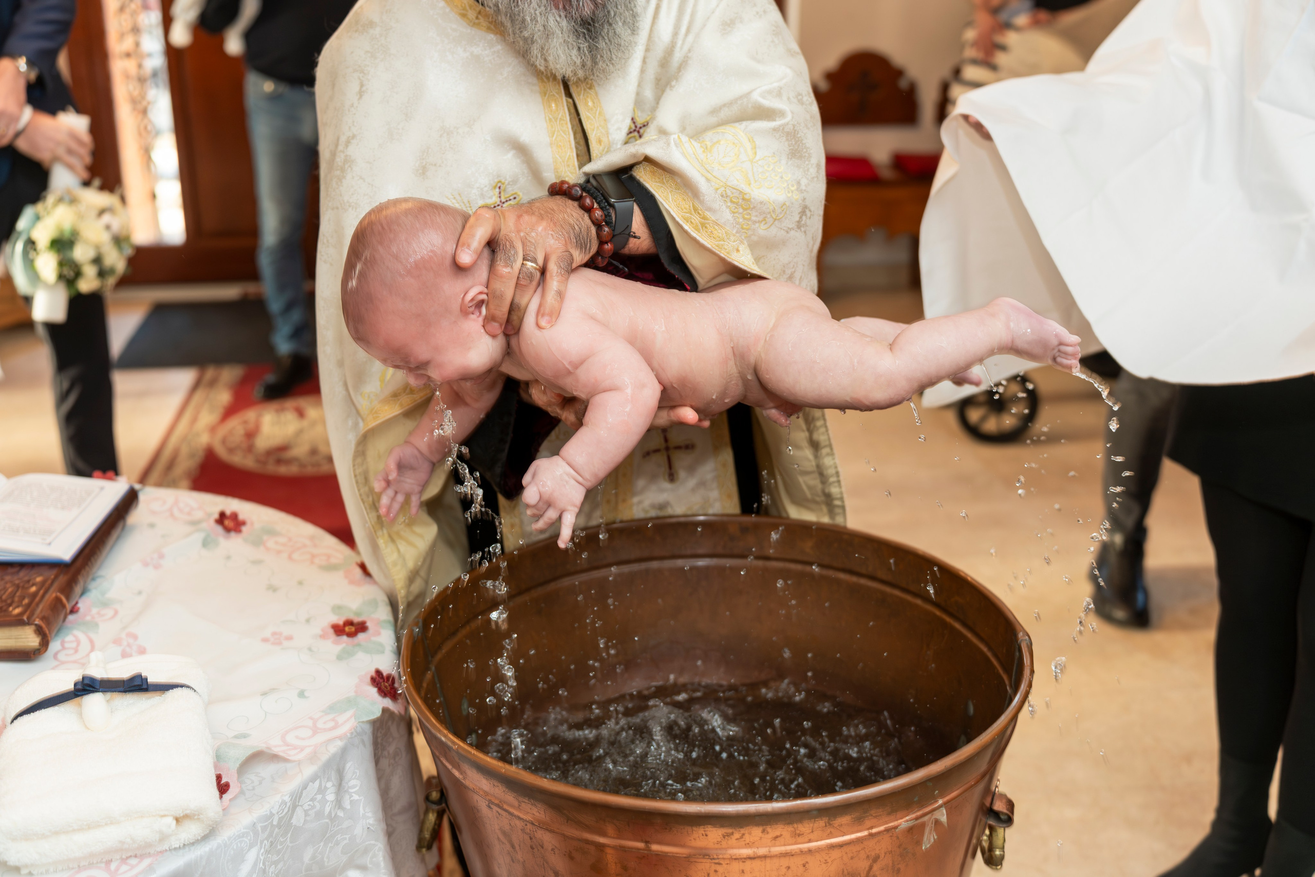 Baby in floral dress held by an adult woman during baptism ceremony with candle and decorations nearby.