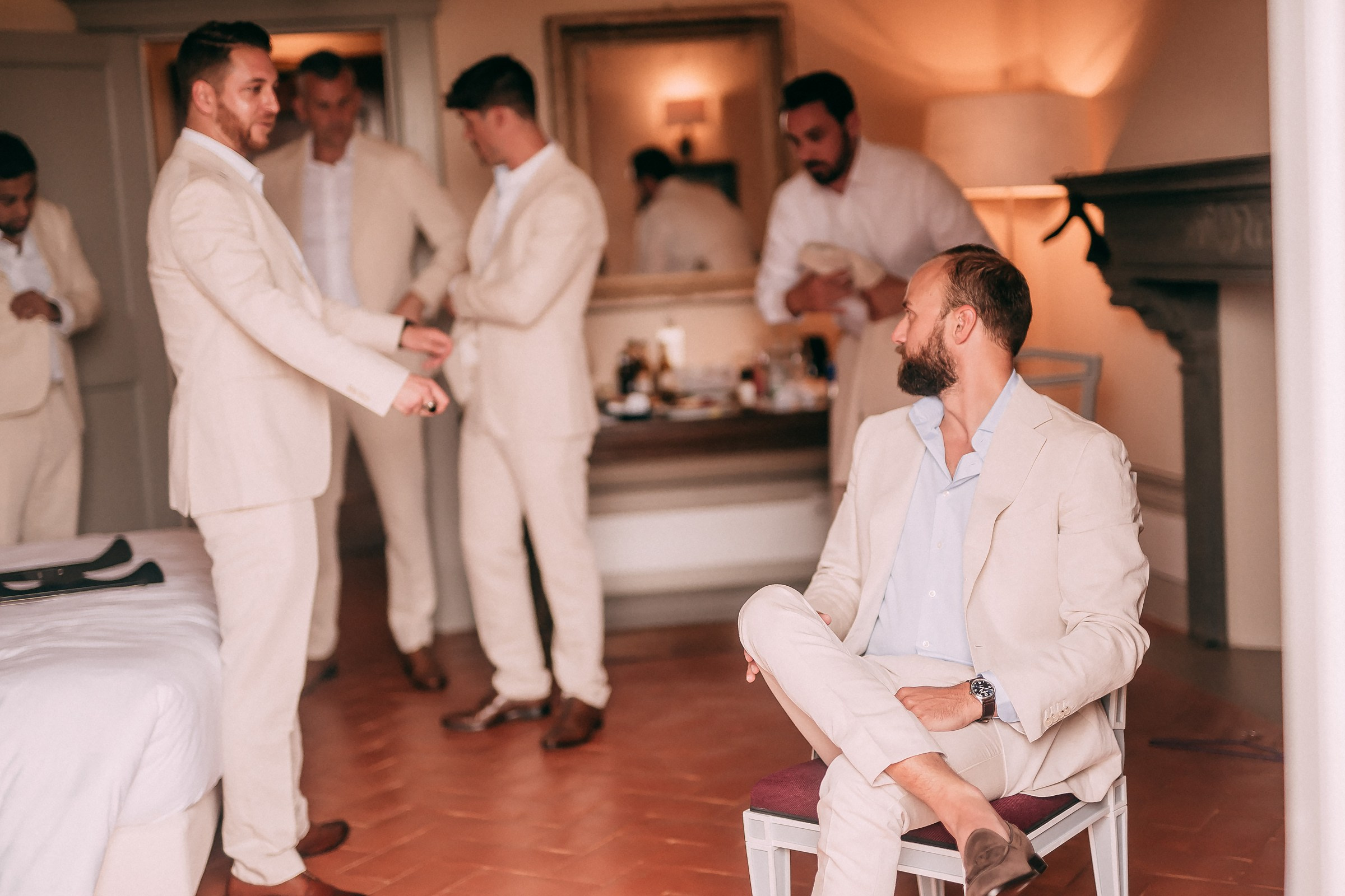The groom calmly seated, dressed in a cream suit, as groomsmen prepare in the background.
