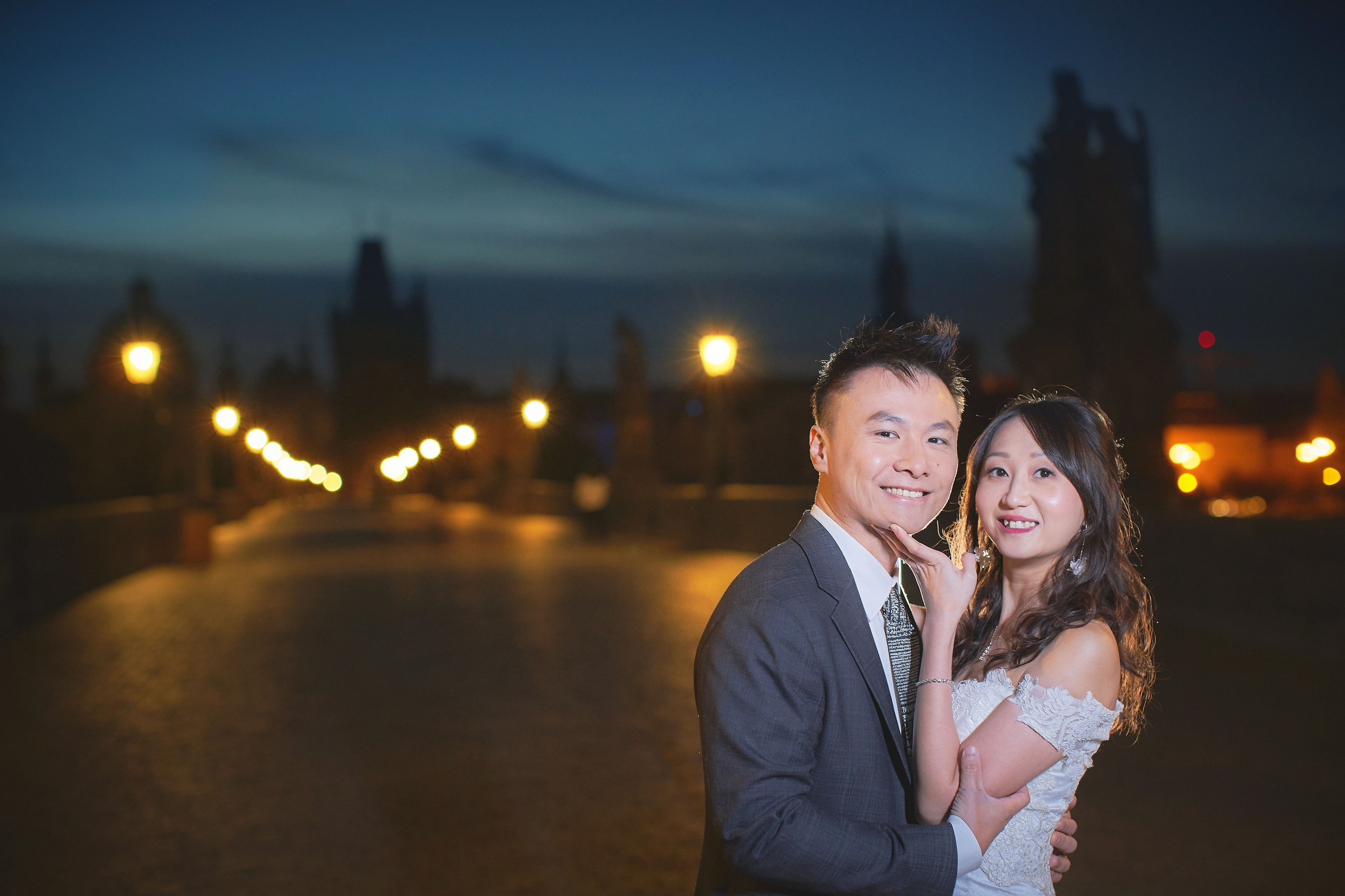 Bride Eva caressing groom Conan’s face while both look confidently at the camera on Charles Bridge, Prague.