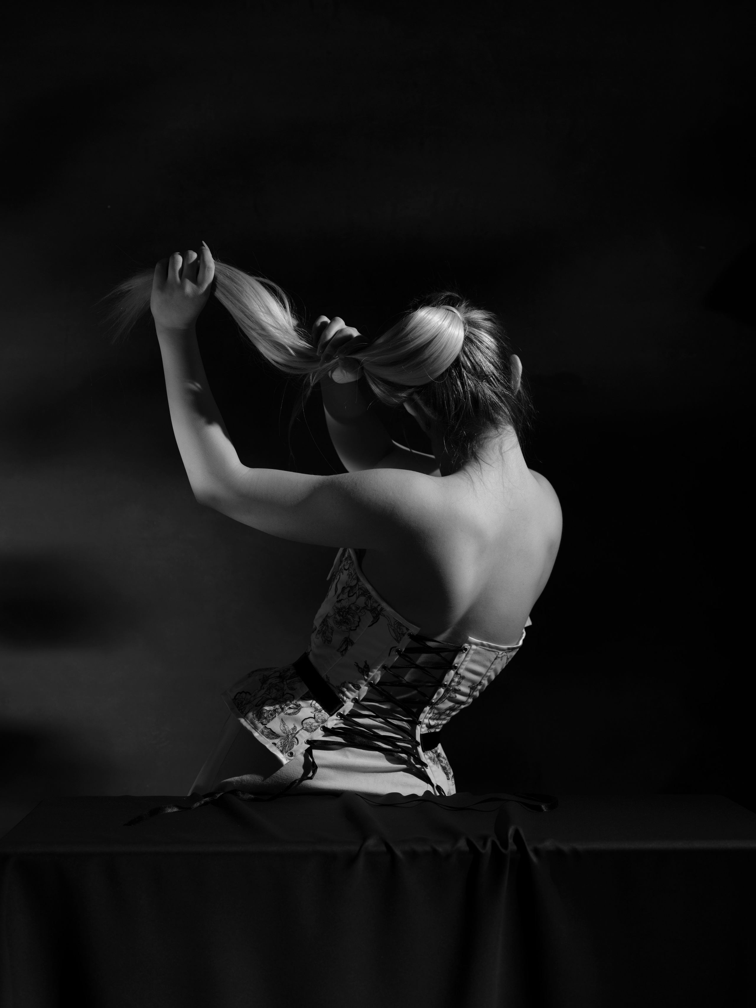 Black and white studio portrait of a woman wearing a corset, seated on a table with her back to the camera, inspired by classic fashion photography.