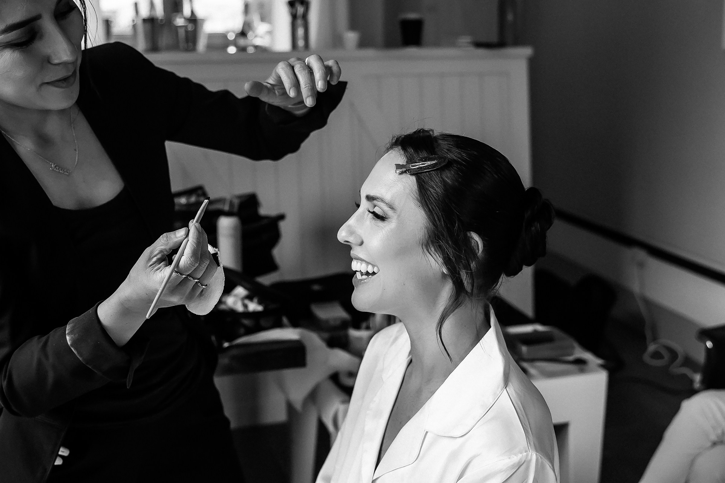 The bride laughing joyfully while her makeup artist finalizes her look, radiating happiness and excitement for her special day.