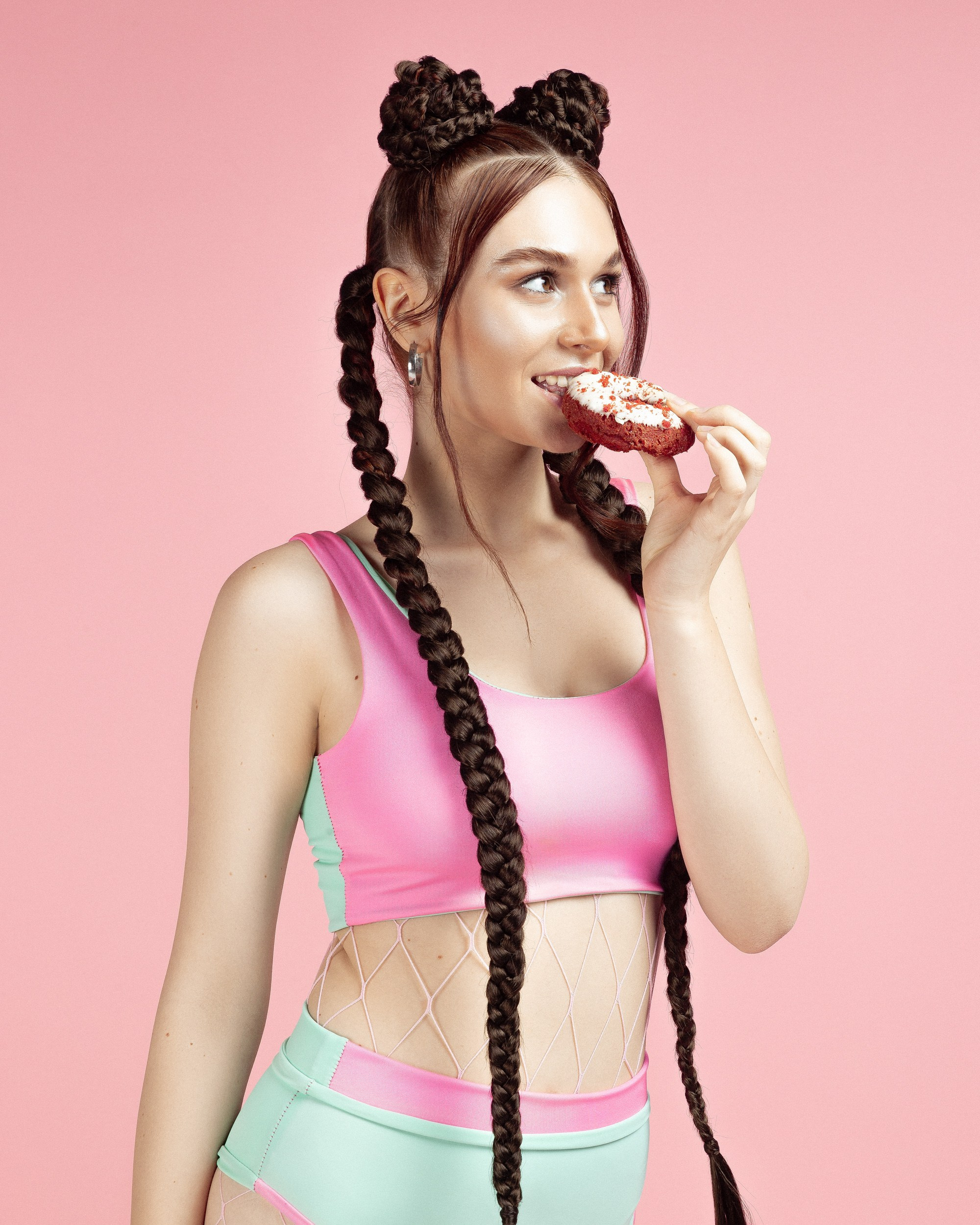 Woman with long braids and fitness wear eating a donut, set against a bright pink backdrop