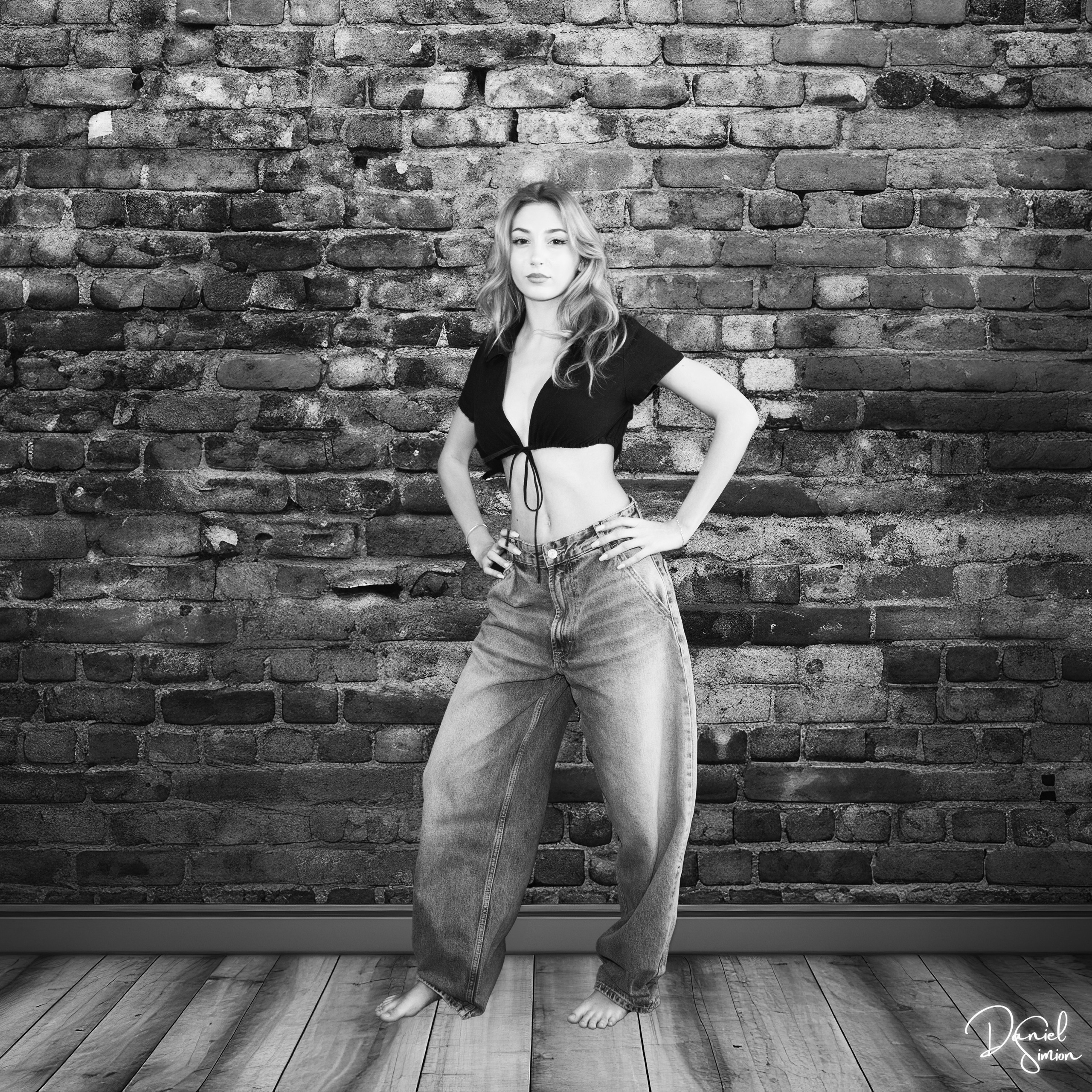 Young woman standing barefoot in front of a brick wall wearing wide jeans and a cropped top.