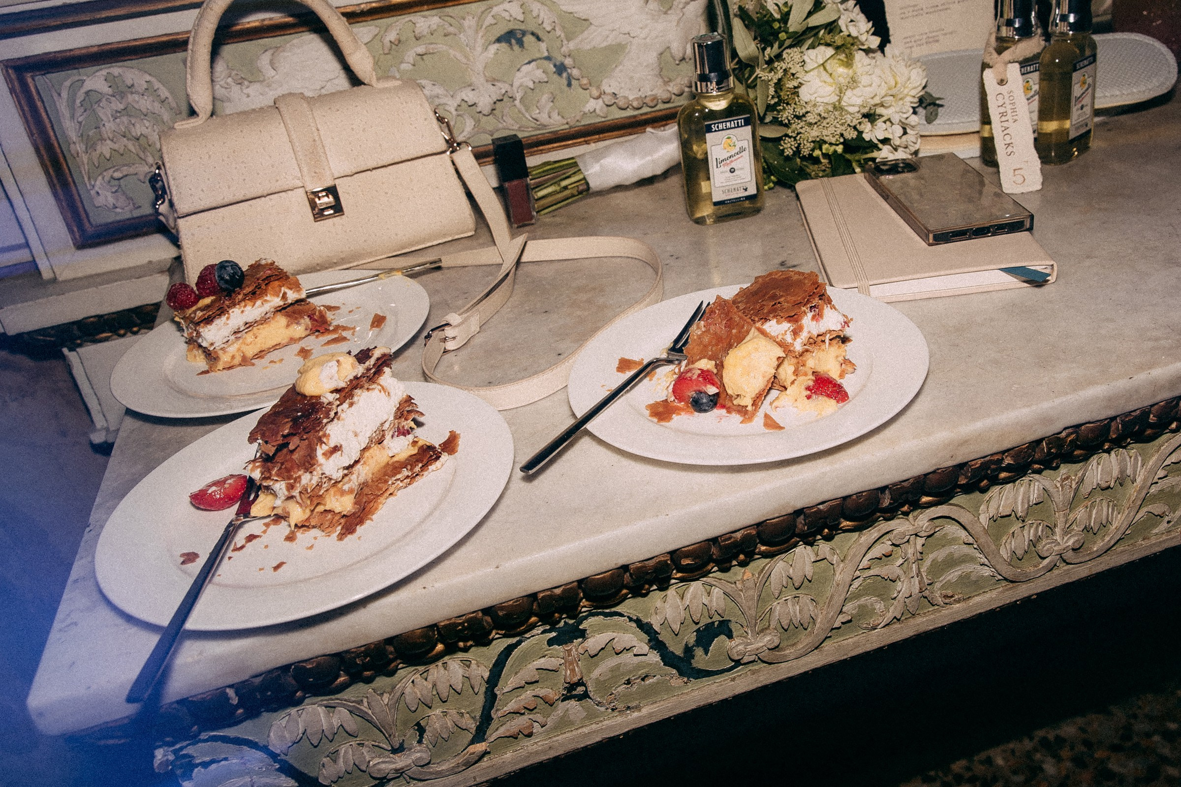 Three partially eaten slices of layered berry and cream cake rest on ornate marble-topped furniture, surrounded by elegant decor, a beige handbag, floral arrangements, and fragrant bottles, capturing the refined ambiance of a wedding dessert table.