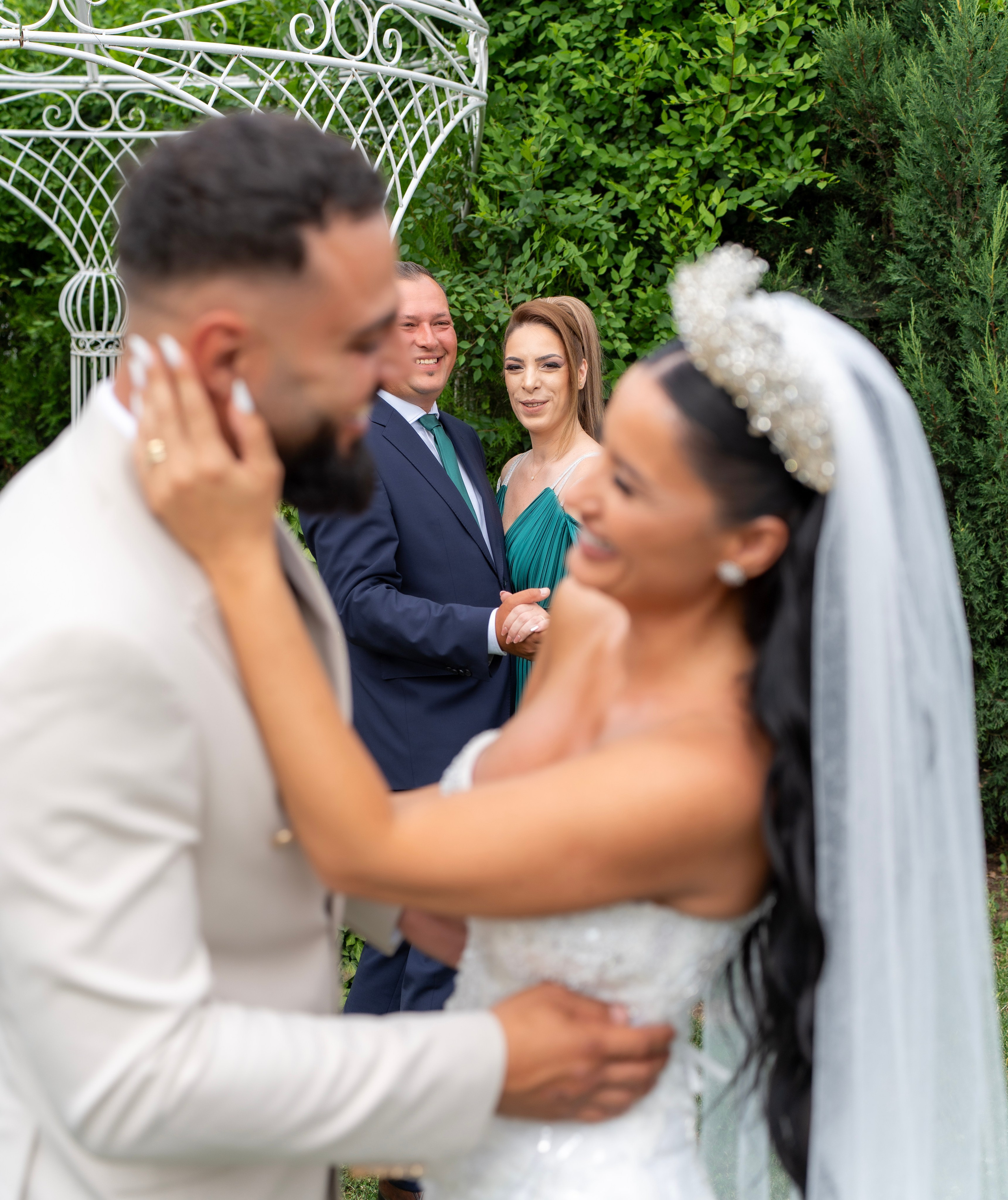 Bride and groom embracing outdoors with two guests smiling behind them near white garden arch and greenery.
