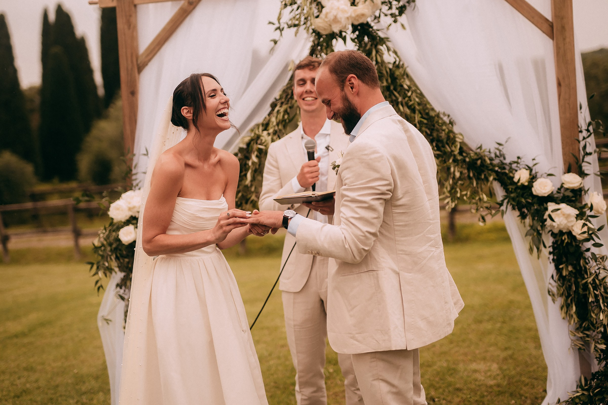 The bride and groom share a joyful laugh during the ring exchange under a beautiful floral arch.
