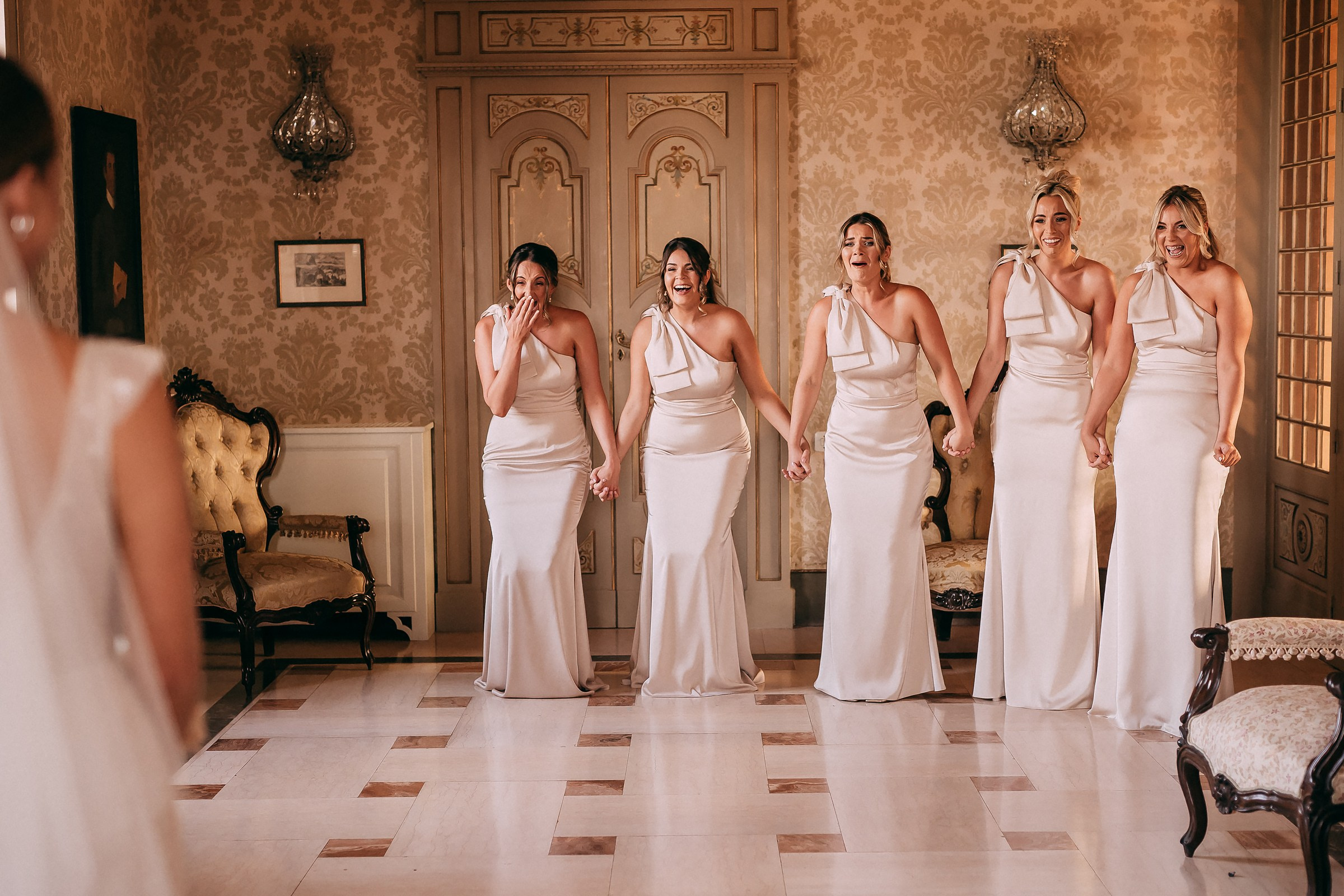 A group of bridesmaids dressed in matching champagne gowns react with heartfelt emotions as the bride makes her grand reveal in the ornate room.
