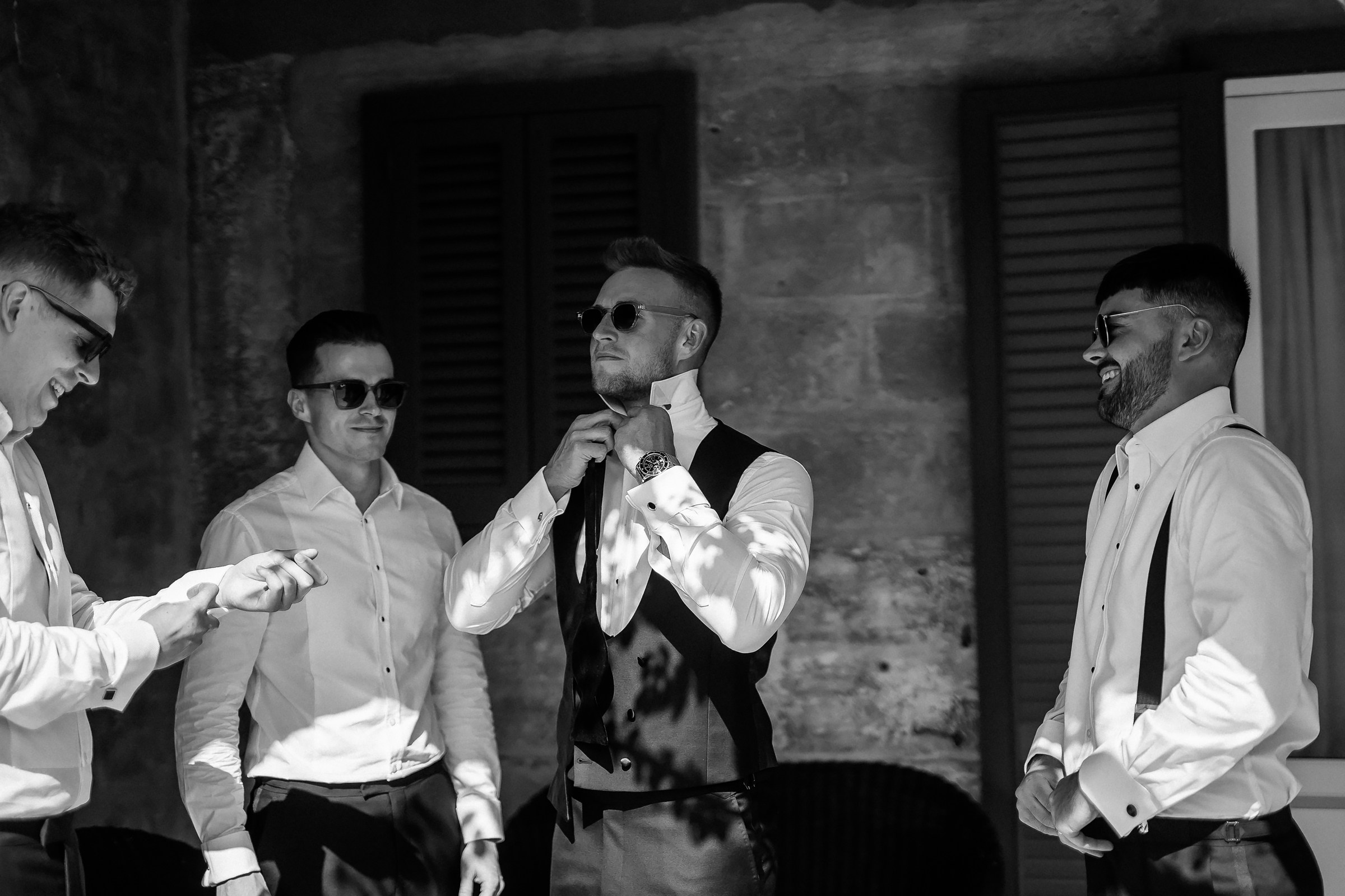 A black-and-white candid shot of the groomsmen, with one adjusting his tie, enjoying the sunny morning outdoors at the historic Masseria Salamina.