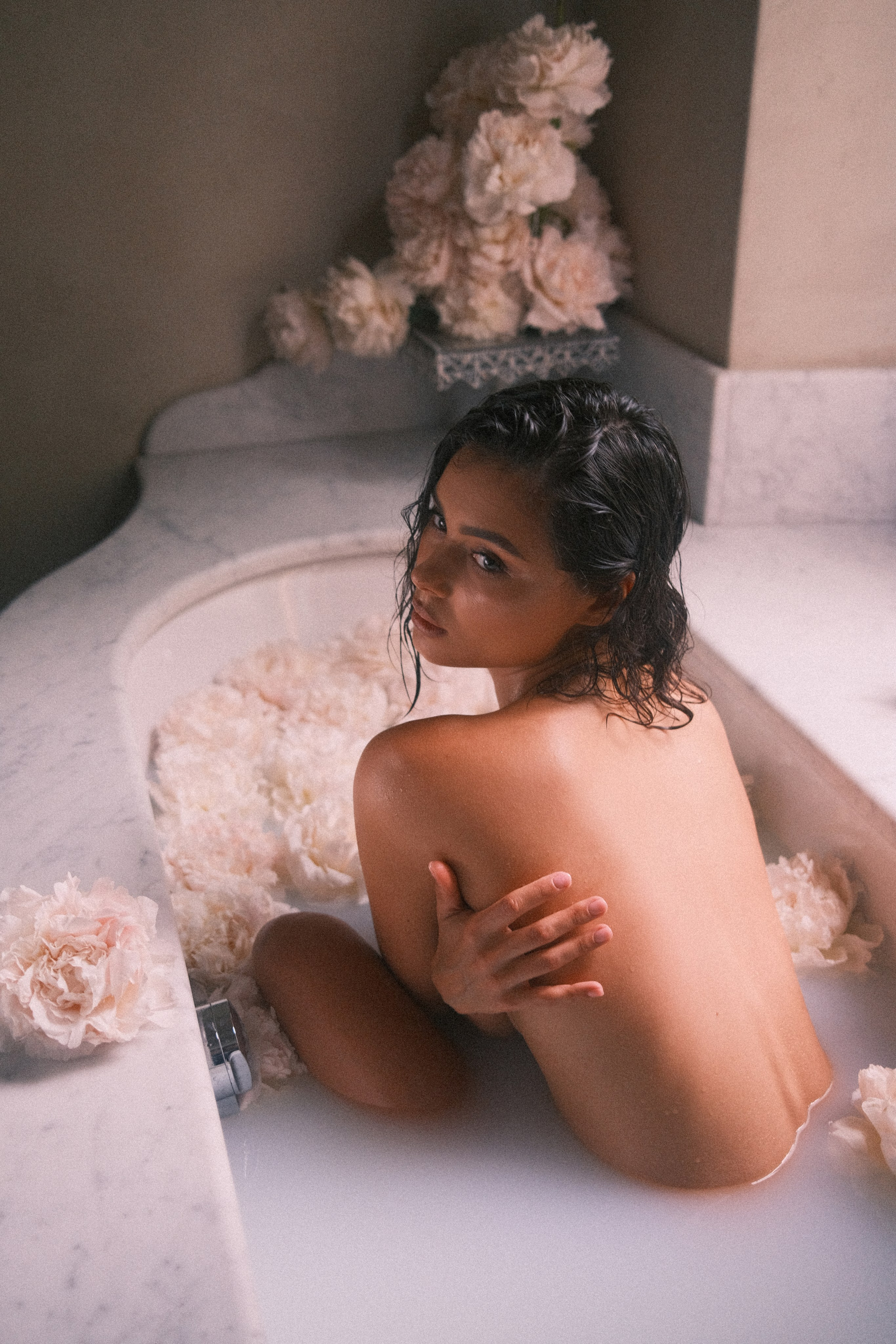 The image captures the bride sitting with her back slightly turned, her pose accentuating elegance while framed by delicate floral arrangements.