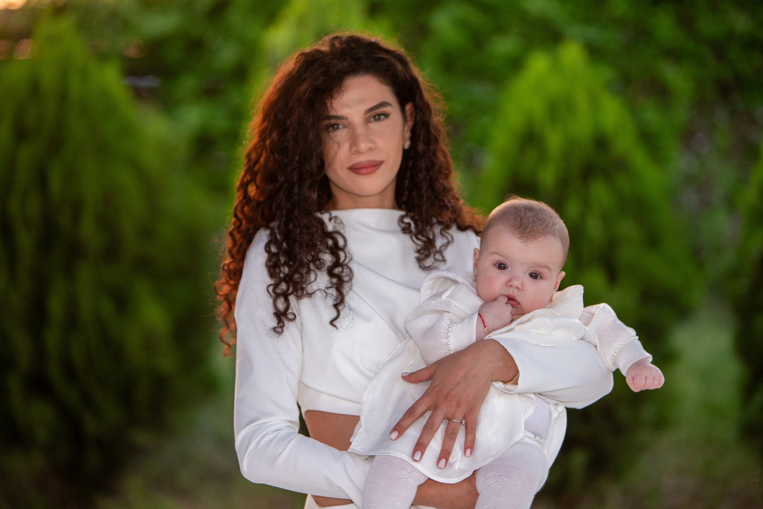 Woman holding a baby dressed in white outdoors with blurred green foliage background.