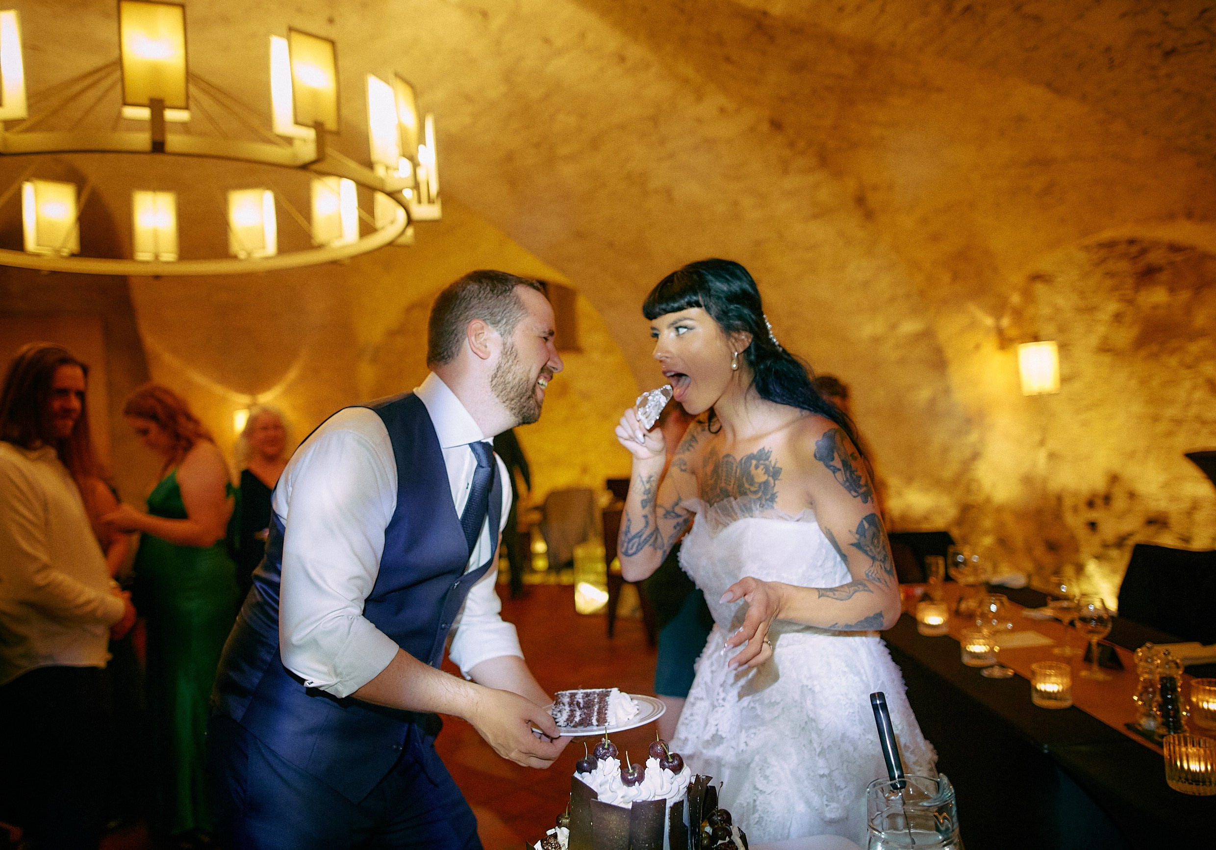 Tattooed bride teasingly licking cake knife with amused groom.
