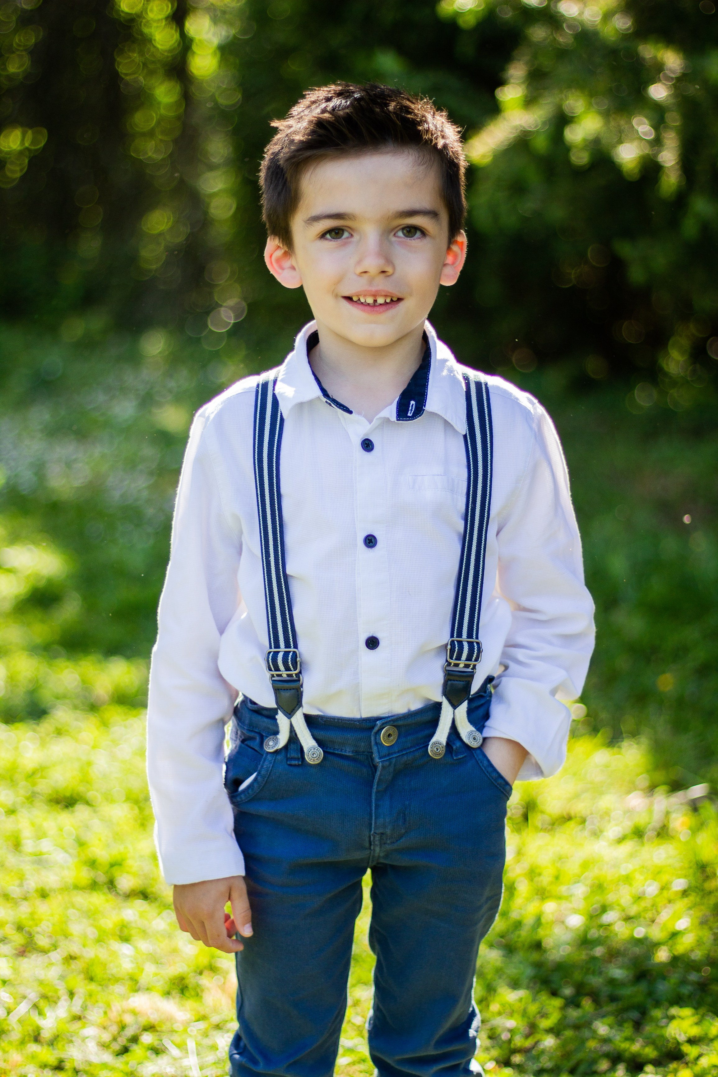 Jeune garçon souriant dans un parc à Poitiers, habillé d'une chemise blanche et de bretelles bleues, posant sous la lumière du soleil avec un fond verdoyant.