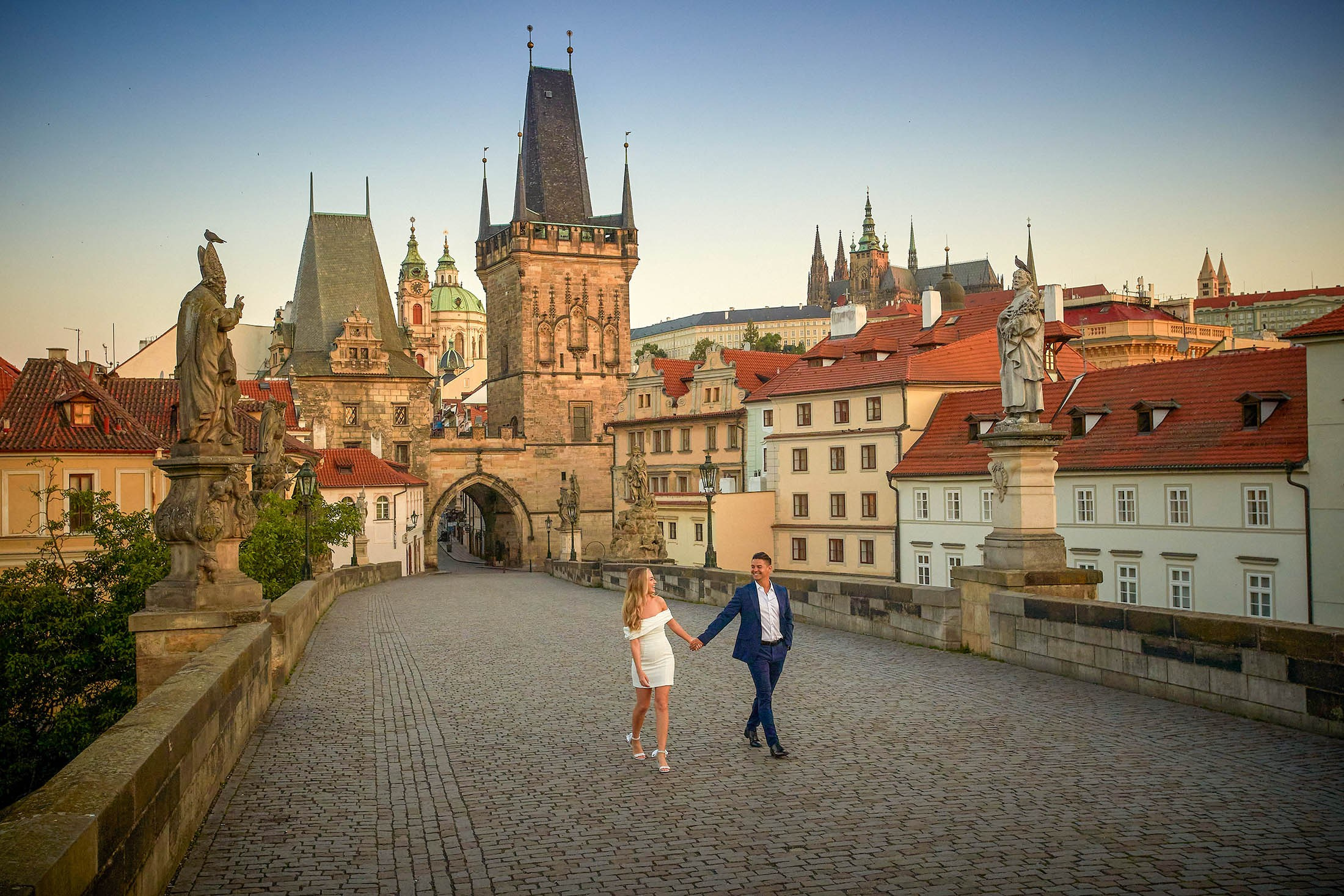A California couple strolling hand-in-hand on empty Charles Bridge in warm sunrise light.