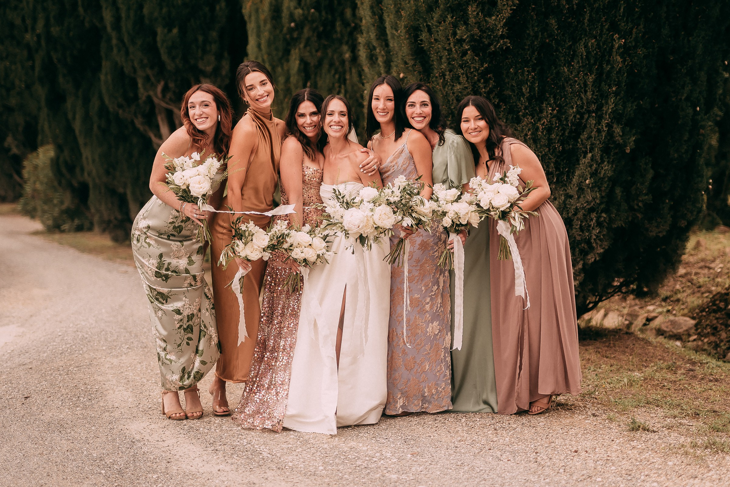 Bride and bridesmaids posing together on a tree-lined pathway, each holding bouquets of white flowers.