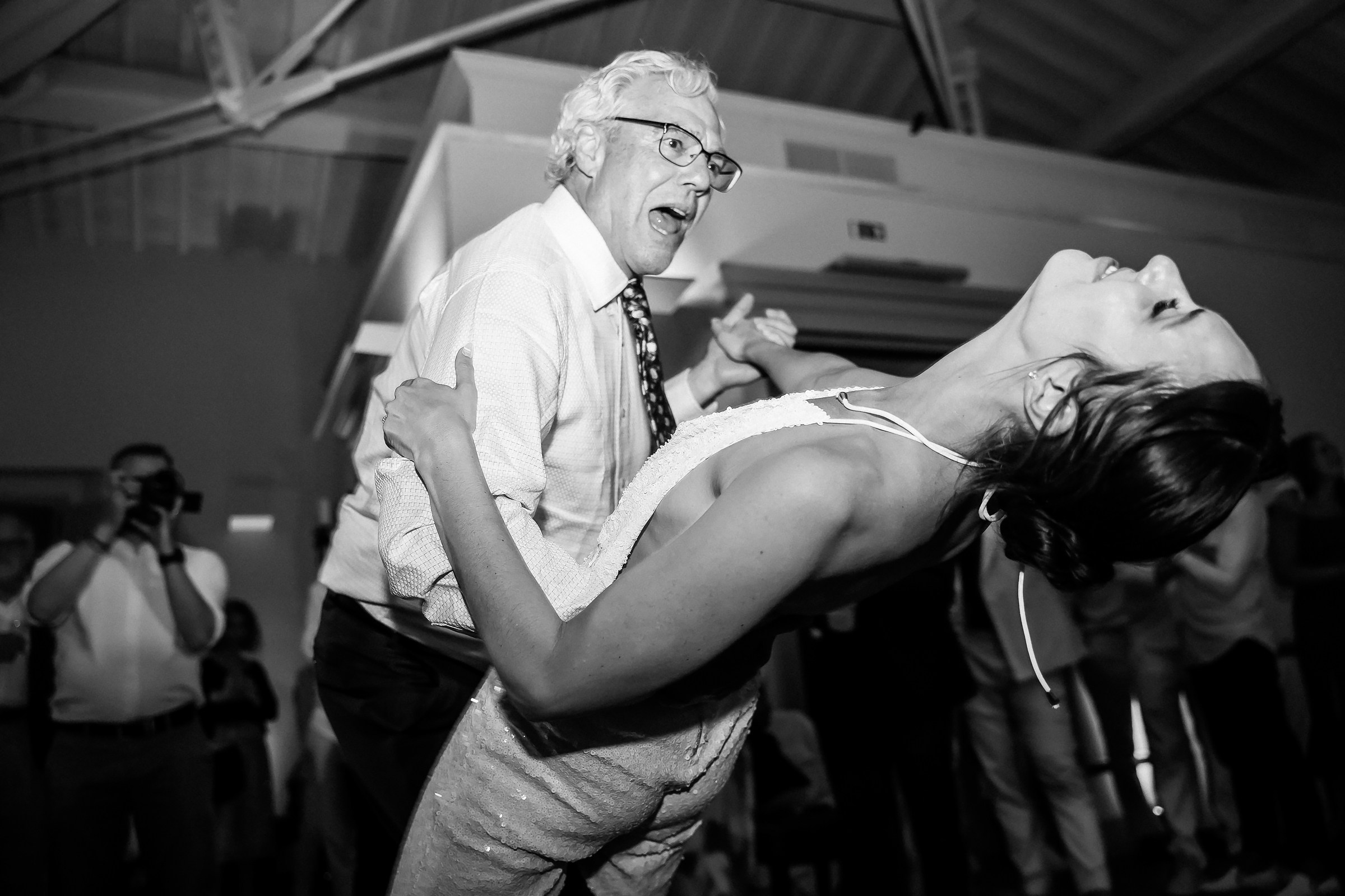 The bride and her father joyfully dip into a dramatic dance move, capturing a moment of laughter and celebration in a black-and-white photograph.