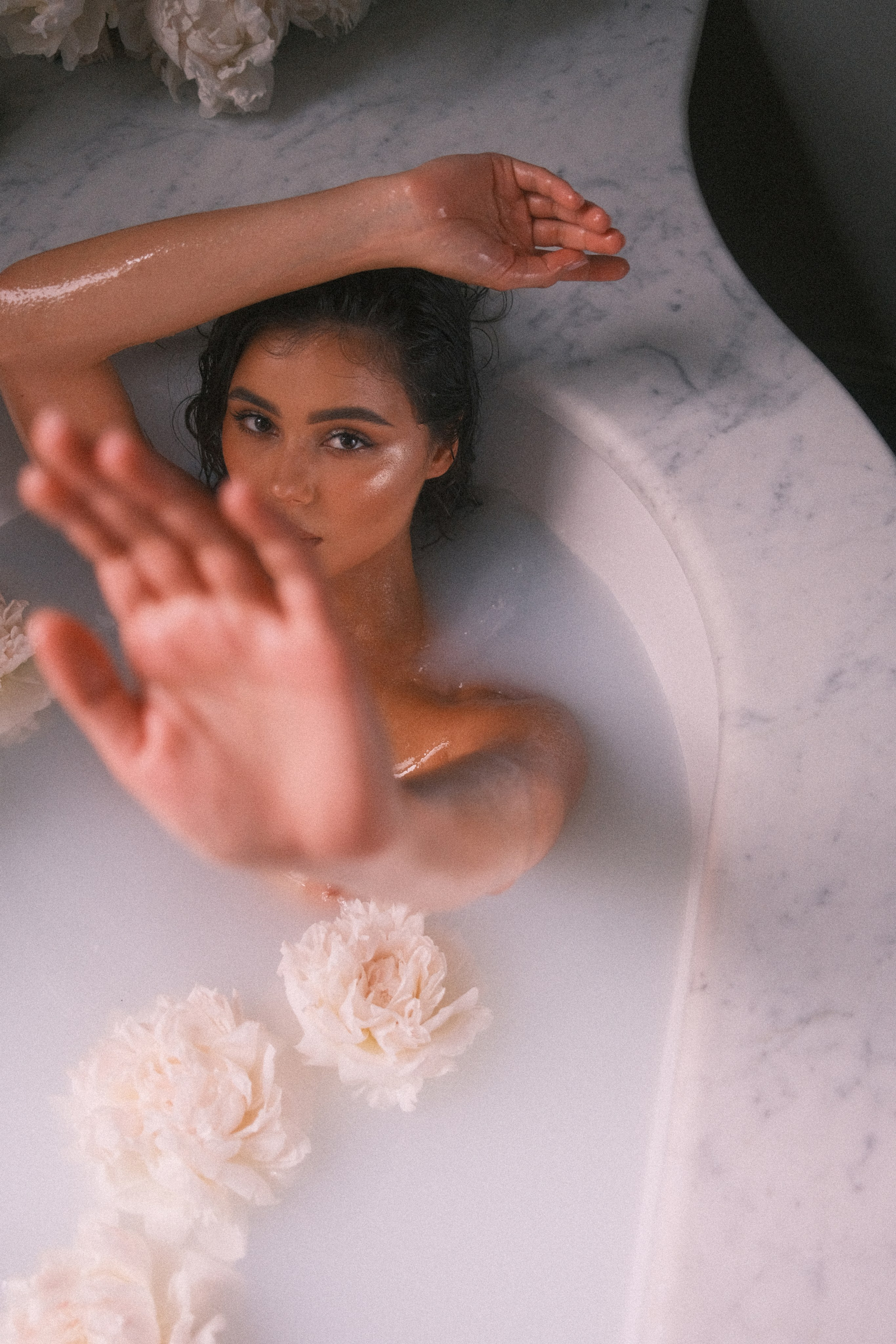 A relaxed and joyful pose of a bride reclining in a bath with a hand raised, amidst a sea of flowers.