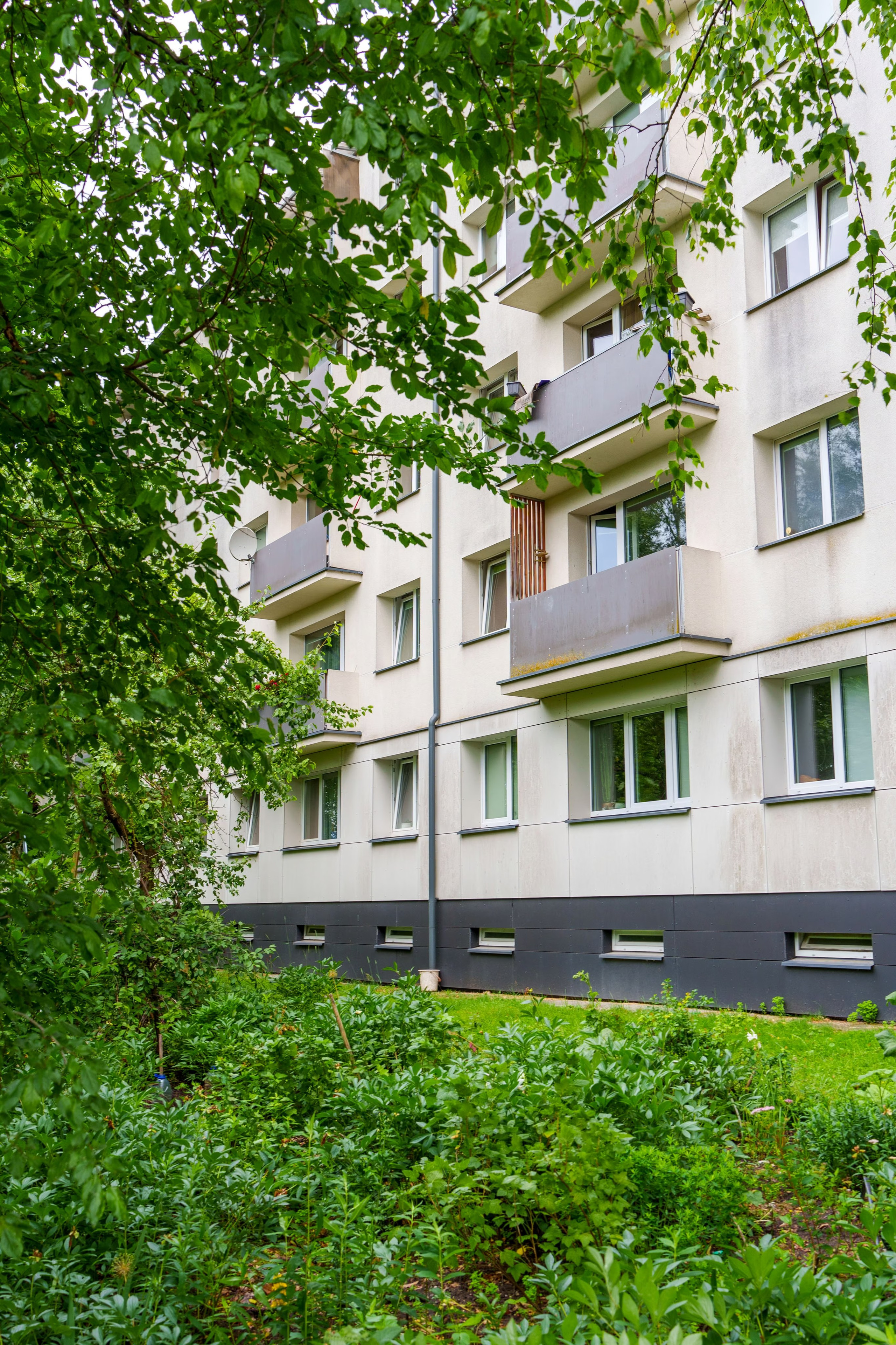 Šiuolaikiško daugiabučio fasadas su balkonais, apsuptas žalumos ir medžių.