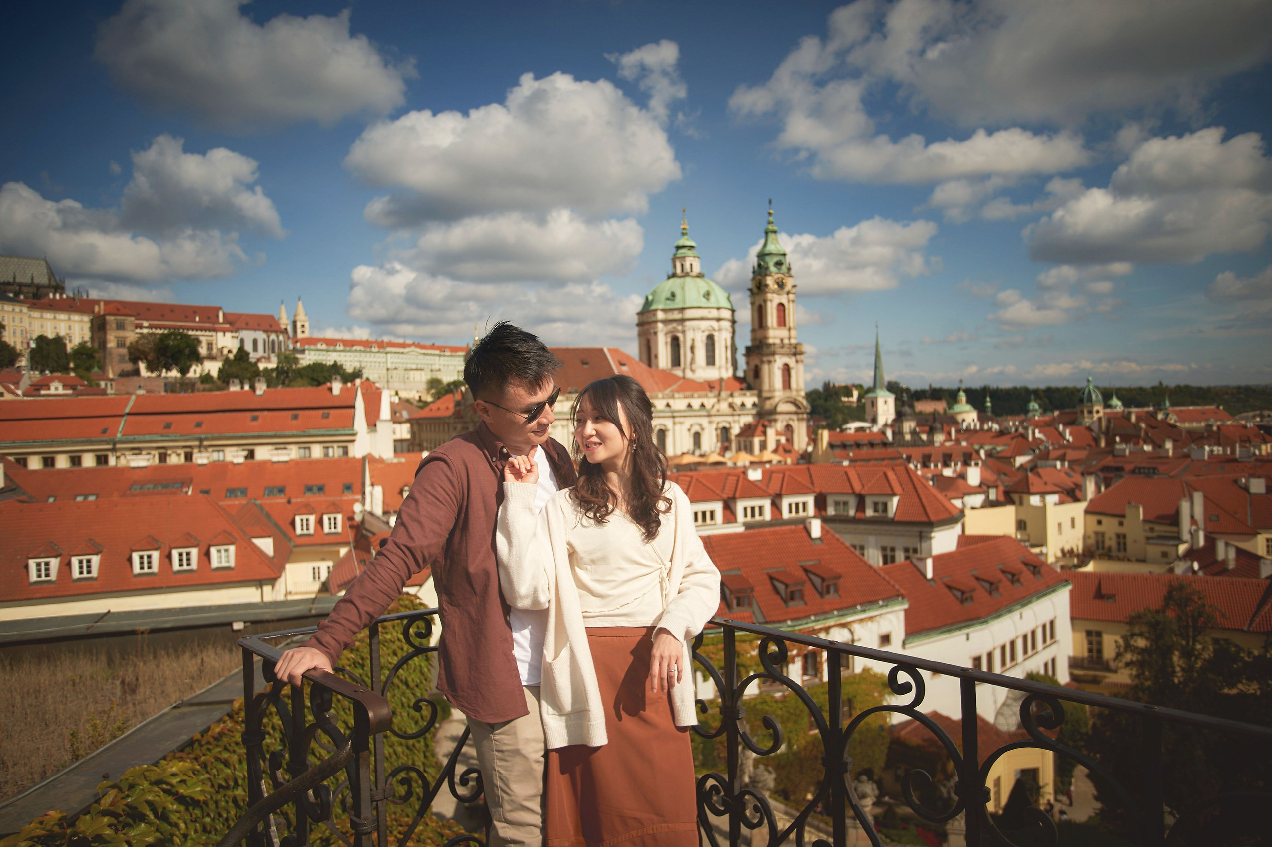 Eva and Conan in casual wear at the top of Vrtba Garden with St. Nicholas Church and clouds.