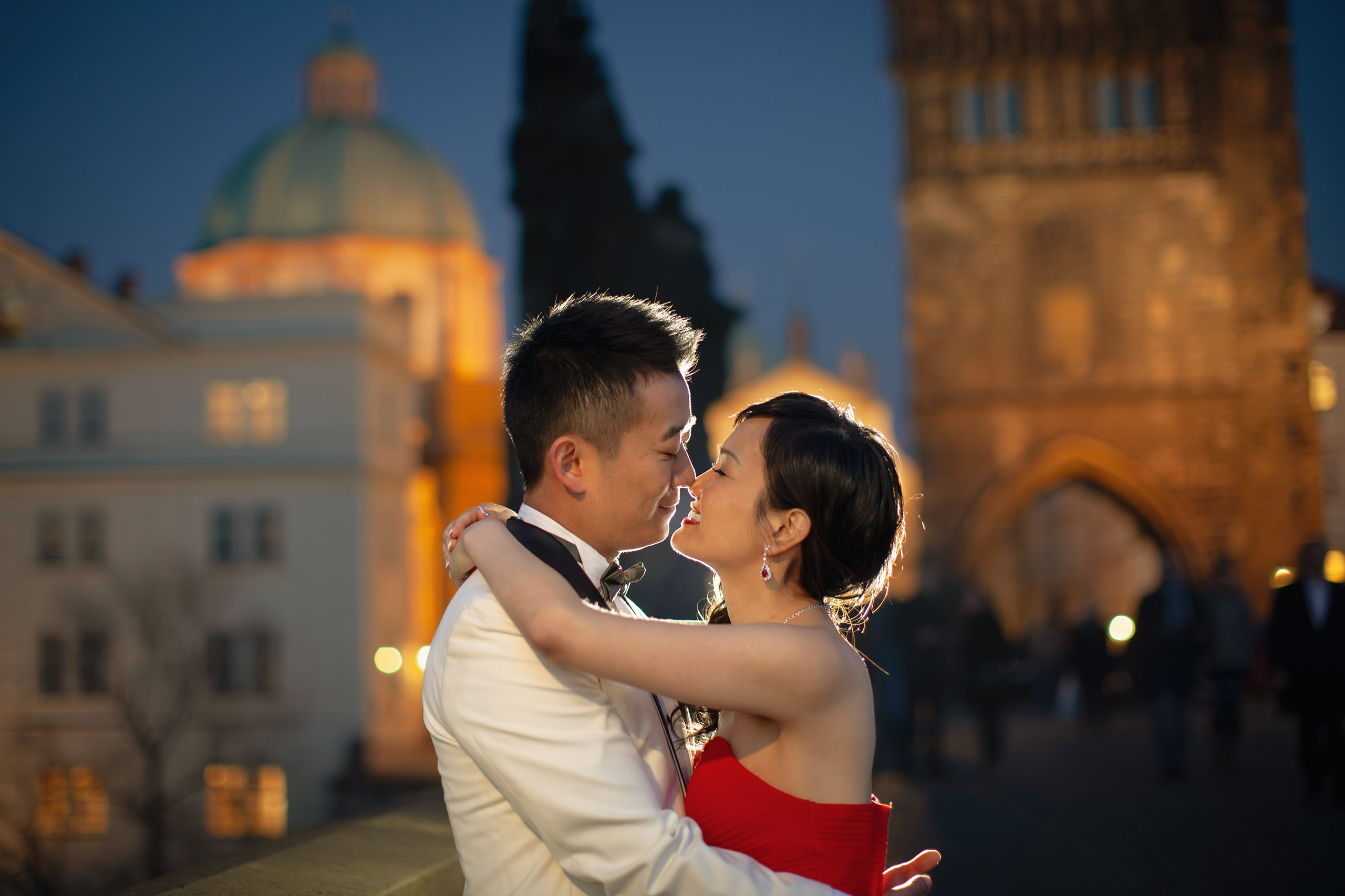 A stylish Hong Kong couple dressed in evening wear snuggle up underneath the historic gas lamps on the Charles Bridge at dusk.