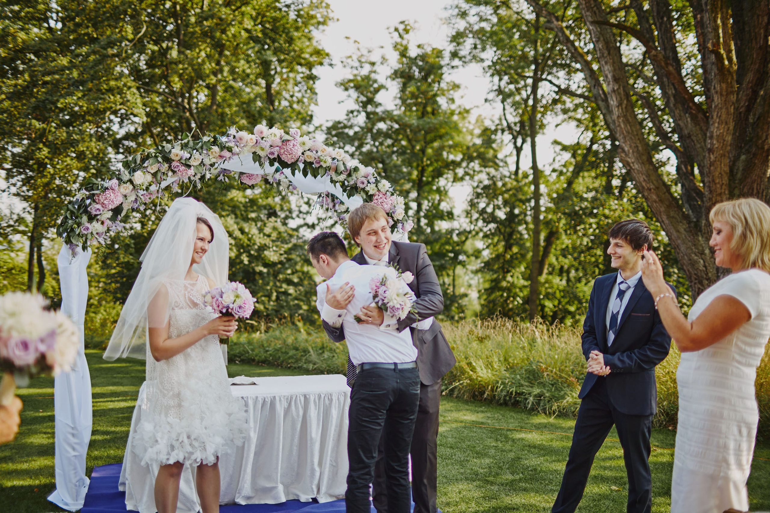 As guests take pictures, a groom and one of his best friends warmly embrace near the floral archway following an outdoor, summer wedding.